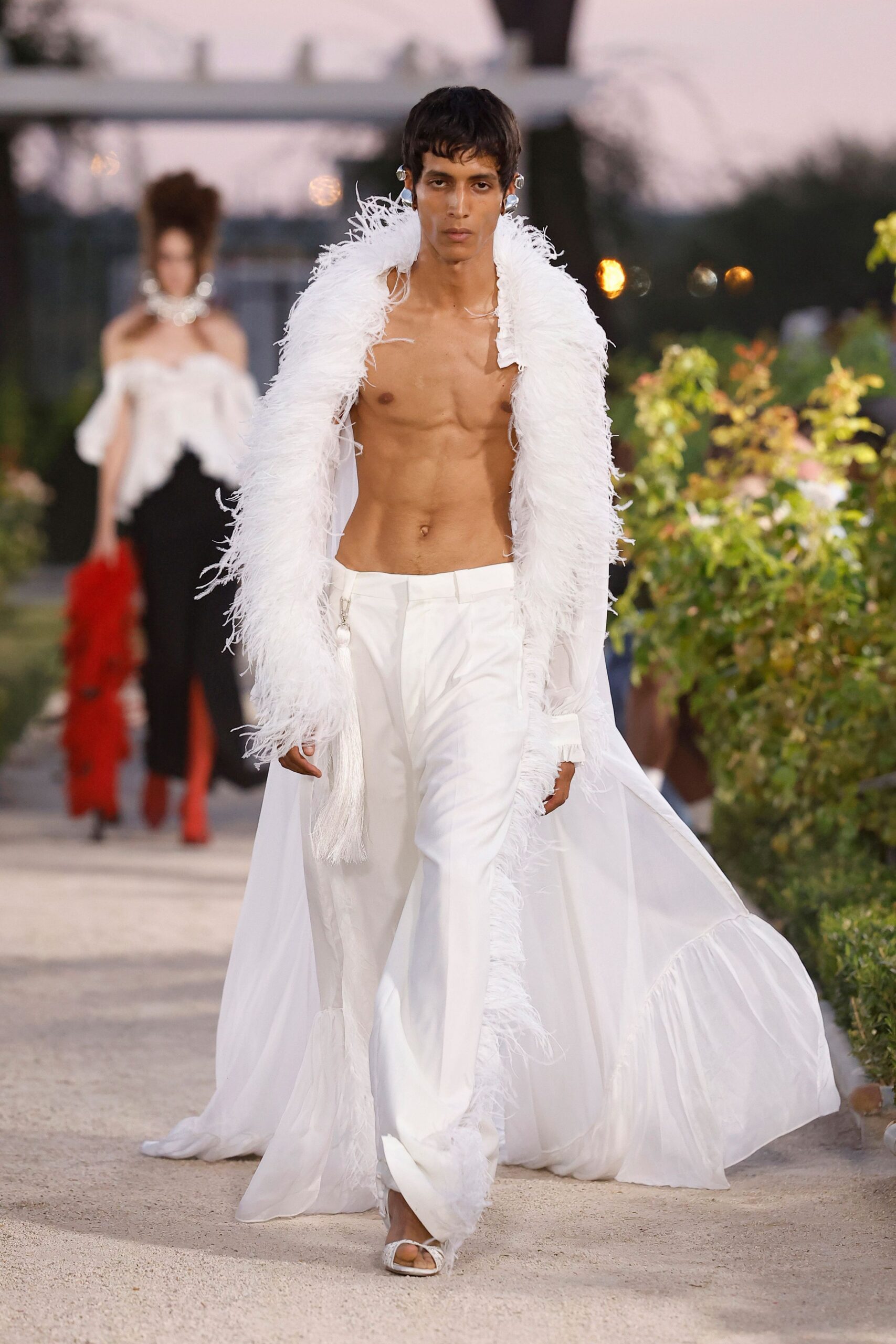 White sheer robe edged in shaggy feather trim, ruffle pooling behind. White low-slung trousers with a zip pull hanging at the hip. White thong sandals, square at the toe. Clear bubble-like ear cuffs. Garden aisle at dusk, carrying the afterparty home on bare skin.