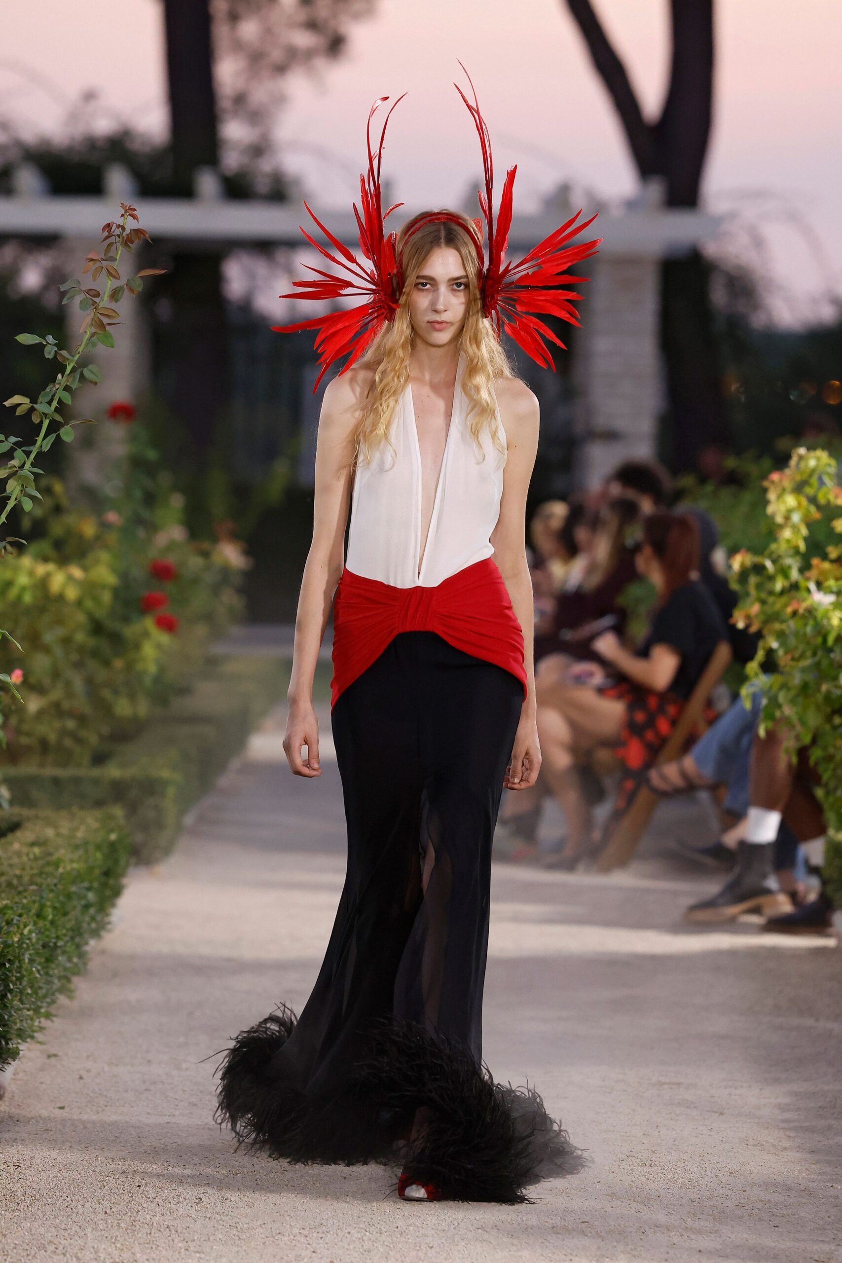 Scarlet feather bursts wired high from a narrow red headband. Ivory plunge bodice meeting a ruched scarlet hip wrap over black chiffon. Black ostrich fluff pooling around the floor-sweeping skirt. Red peep-toe pumps flashing under the sheer front slit. Rose garden at dusk, when the party has turned slightly feral.