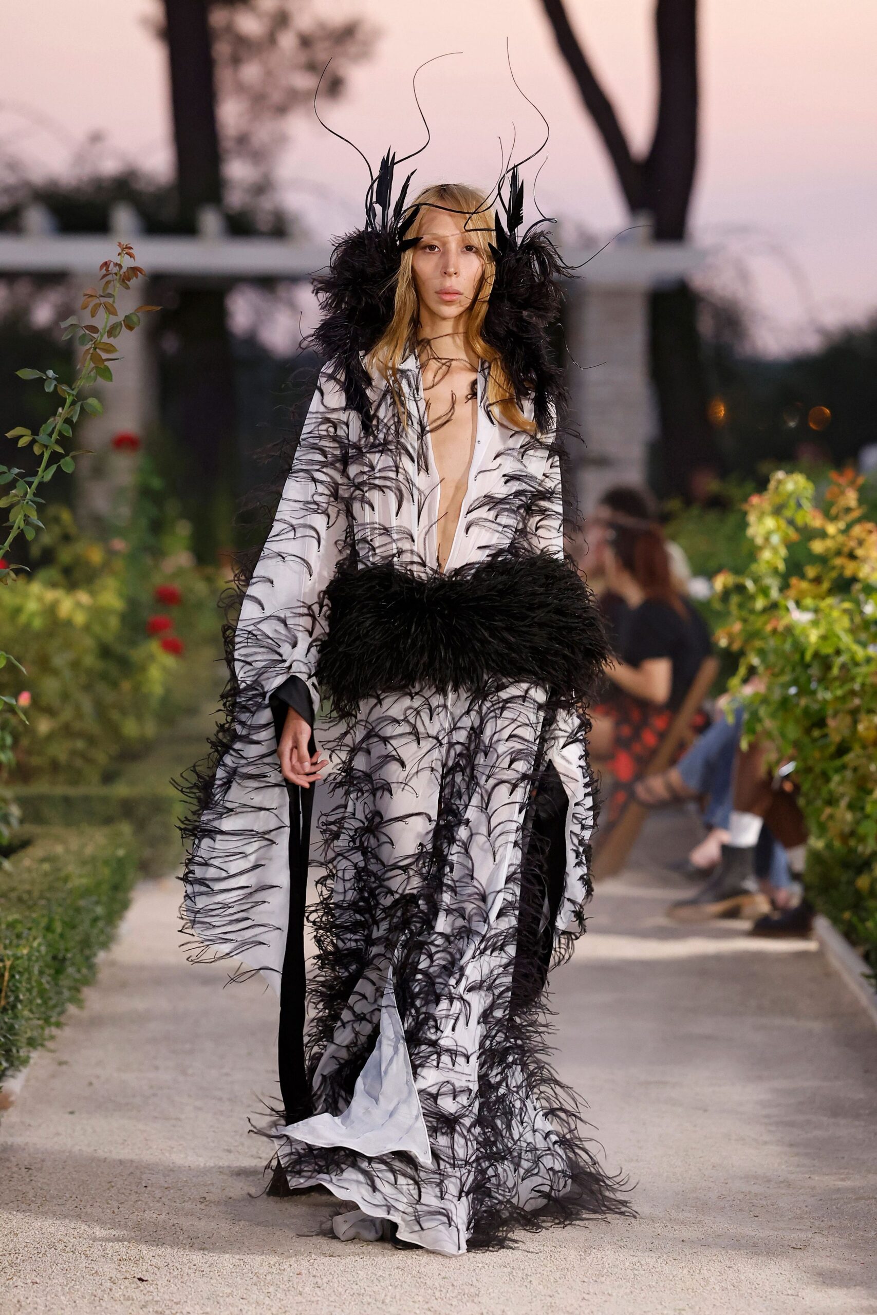 Black feather halo with wire tendrils rising above the forehead. White floor-length gown, plunge front and sleeves overgrown with black ostrich fronds. Dense black feather muff cinched low, swallowing the hands. Rose garden at dusk, when the air cools faster than the stone path.