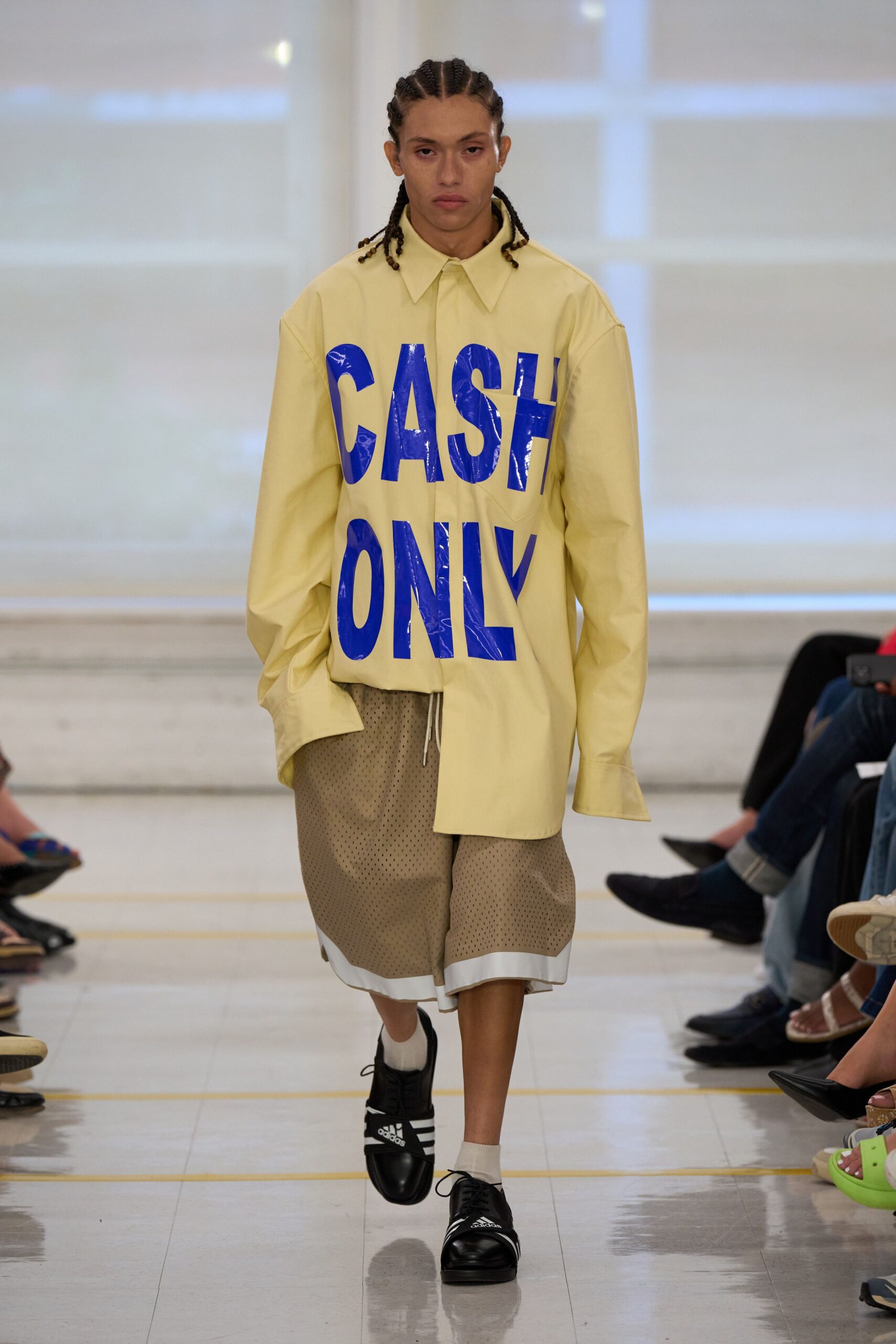 Pale butter shirting blown oversized, covered in cracked cobalt block letters. Taupe mesh basketball shorts with a white underlayer peeking below. Cream crew socks scrunched at the ankle. Black leather trainers with white side stripes and loose laces. Walking to the corner shop in borrowed kit after sleeping through noon.