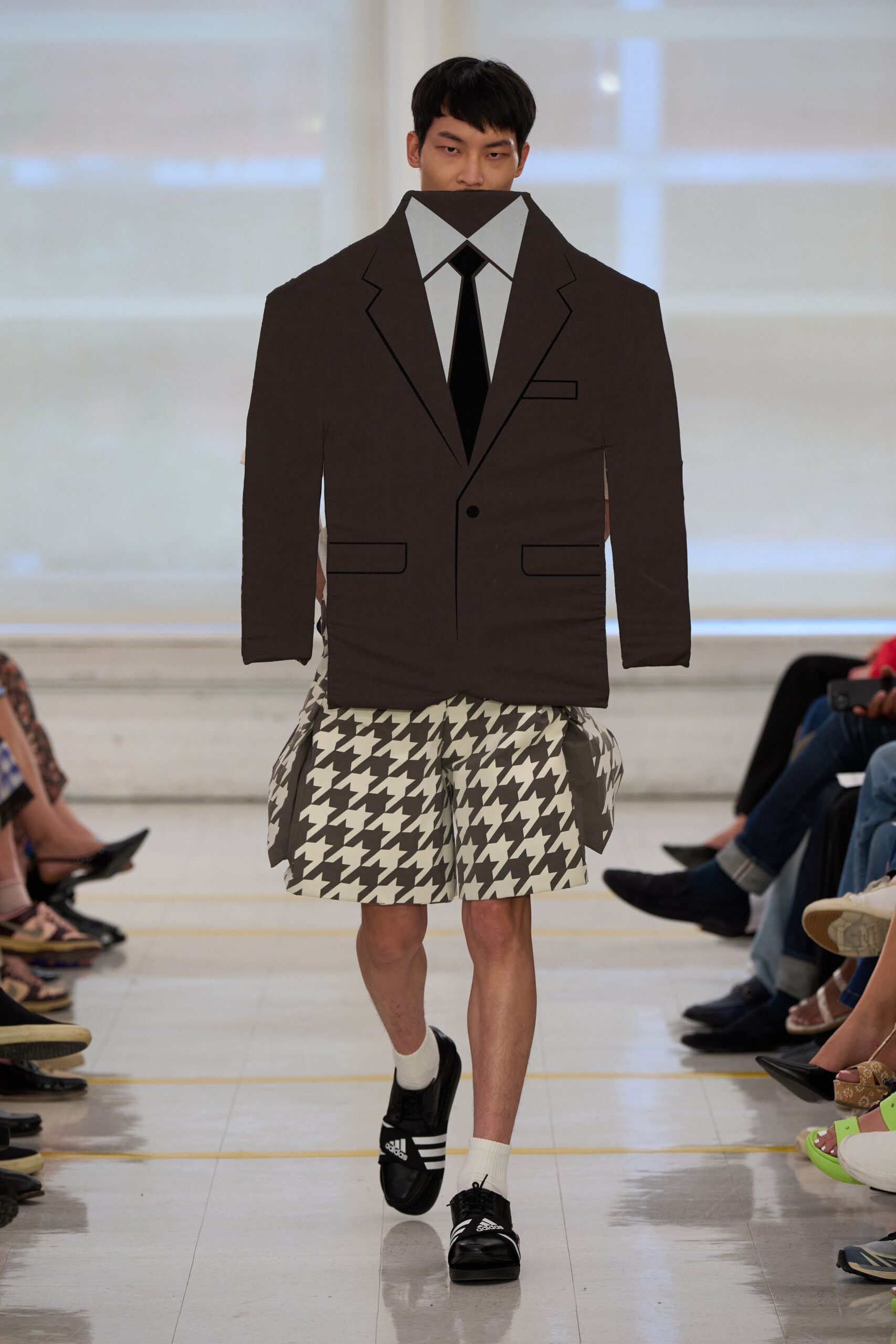 Flat padded cutout blazer with printed shirtfront and narrow black tie. Black-and-white houndstooth shorts, ballooned wide with boxy side panniers. White rib socks. Black sport slides with three white stripes. For crossing the hotel lobby after a pool dip, still wearing half a joke.