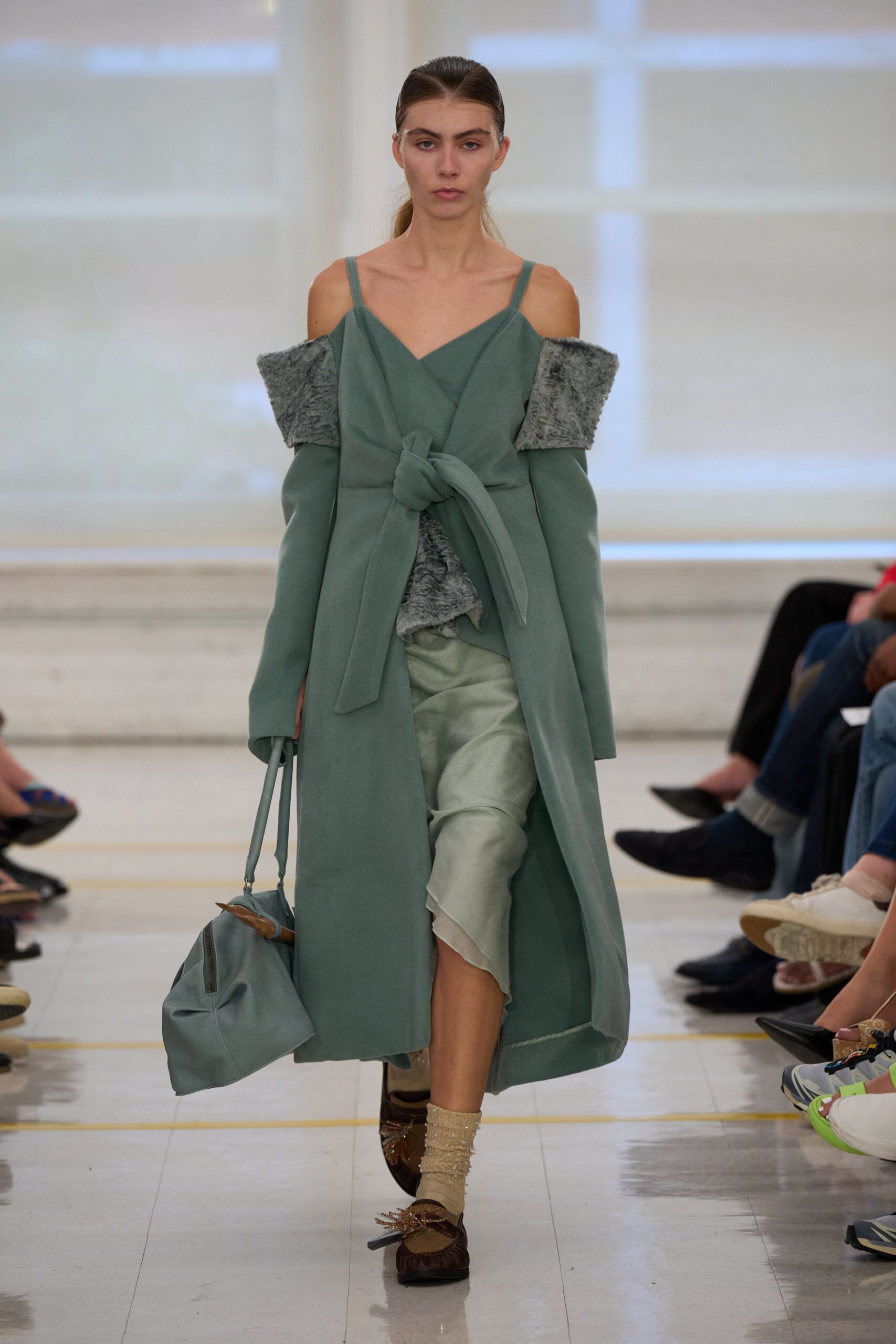 Sage wrap coat, tied in a thick knot over grey faux-fur panels. Pale celadon satin slip, hem hanging in a soft uneven point. Slouchy sage holdall with a wooden clasp peeking out. Beige beaded ankle socks. Brown square-toe flats with tasseled ties. For carrying flowers home through a lobby that still smells of bleach.