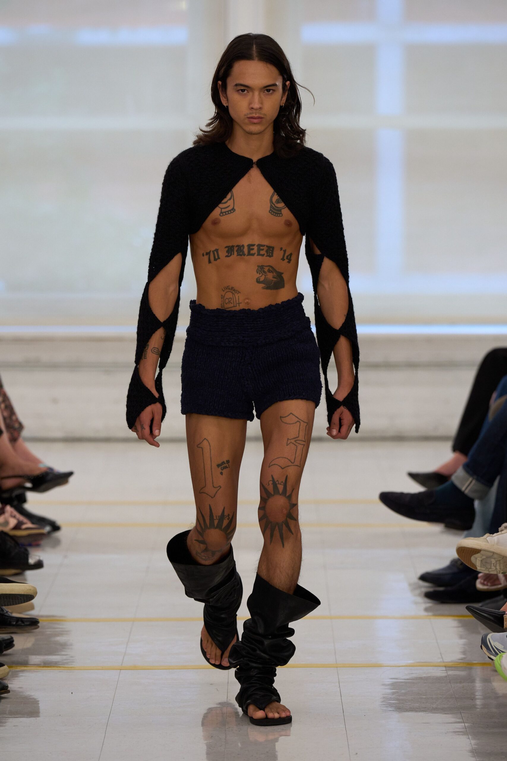 Black boucle knit shrug, hooked once at the throat and split along the sleeves. Navy nubby knit shorts with a thick rolled waistband. Black leather wrap sandals collapsed into slouched tubes around the calves. Whatever you pull on for a dawn swim after not sleeping at all.