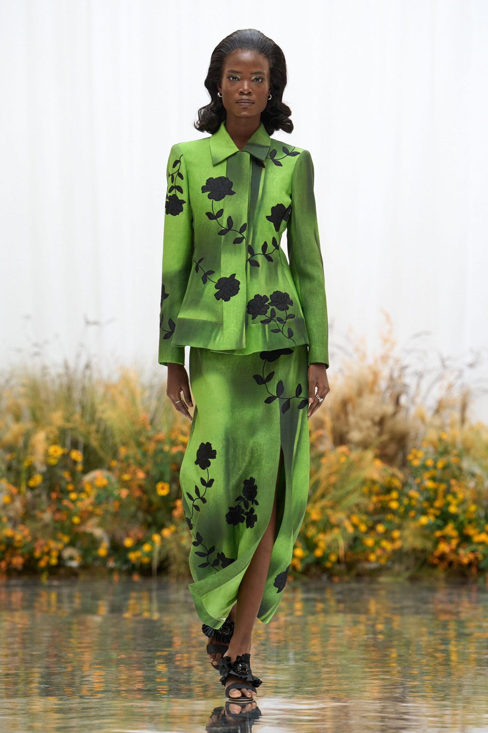 Acid-green tailored jacket with black floral appliqué and a peplum kick. Matching satin skirt, wrapped front opening into a narrow slit. Black ruffled sandals. Crossing the conservatory path after the glasshouse has just been watered.