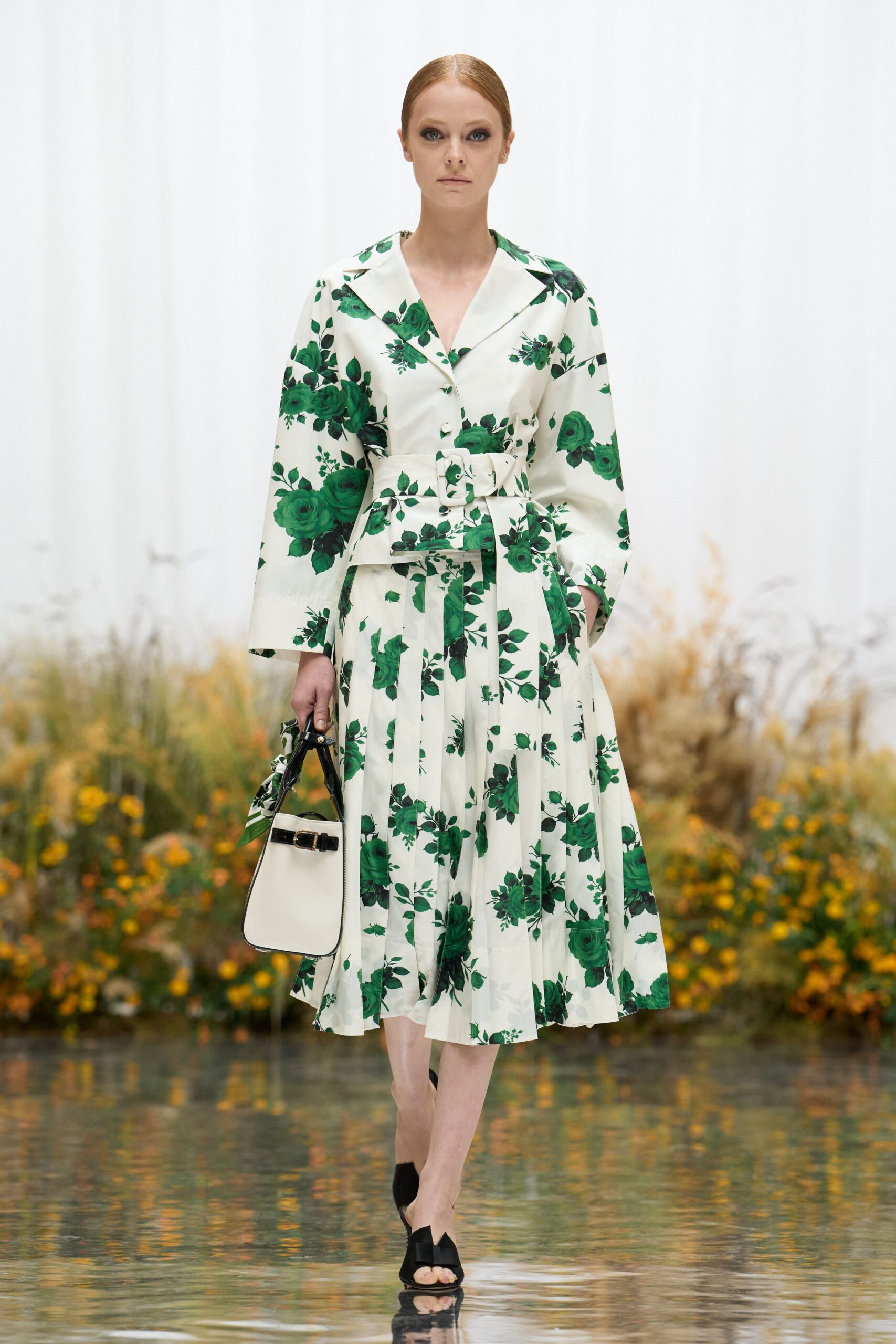 Ivory cotton shirt dress printed with deep green roses, wide notched collar. Matching belt with a covered square buckle cinching the waist. Cream trapeze top-handle bag with black buckle strap and tied scarf. Black slide sandals with a squared vamp. Crossing a conservatory path just after the watering hoses stop.