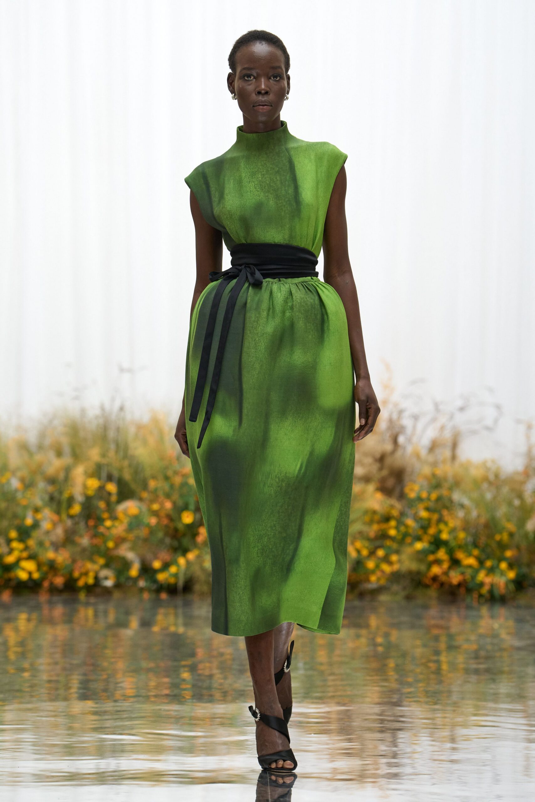 Grass-green satin dress with a high stand collar and cap sleeves. Wide black sash knotted at the waist, one ribbon hanging longer. Black satin ankle-wrap sandals with a small crystal flourish. Crossing a reflecting pool while the garden heat still holds after rain.