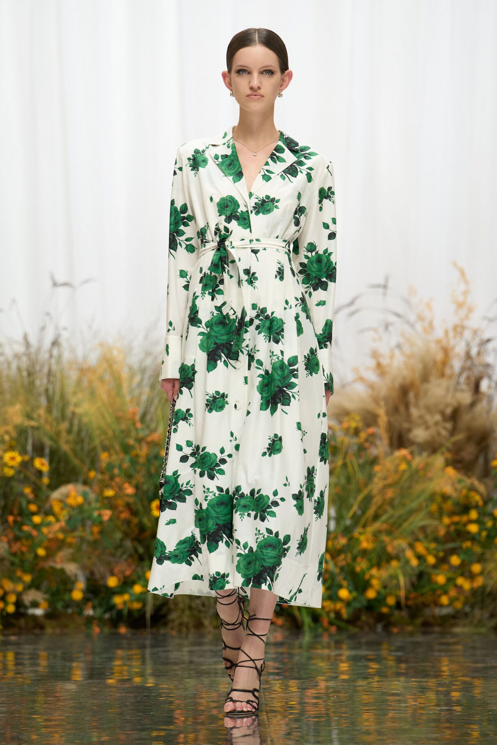 Ivory shirt dress scattered with emerald rose sprays, tied low with a self belt. Small black chain bag hanging straight from one hand. Black lace-up sandals, cords winding nearly to mid-calf. Crossing a hotel garden after rain, trying not to sink into the grass.
