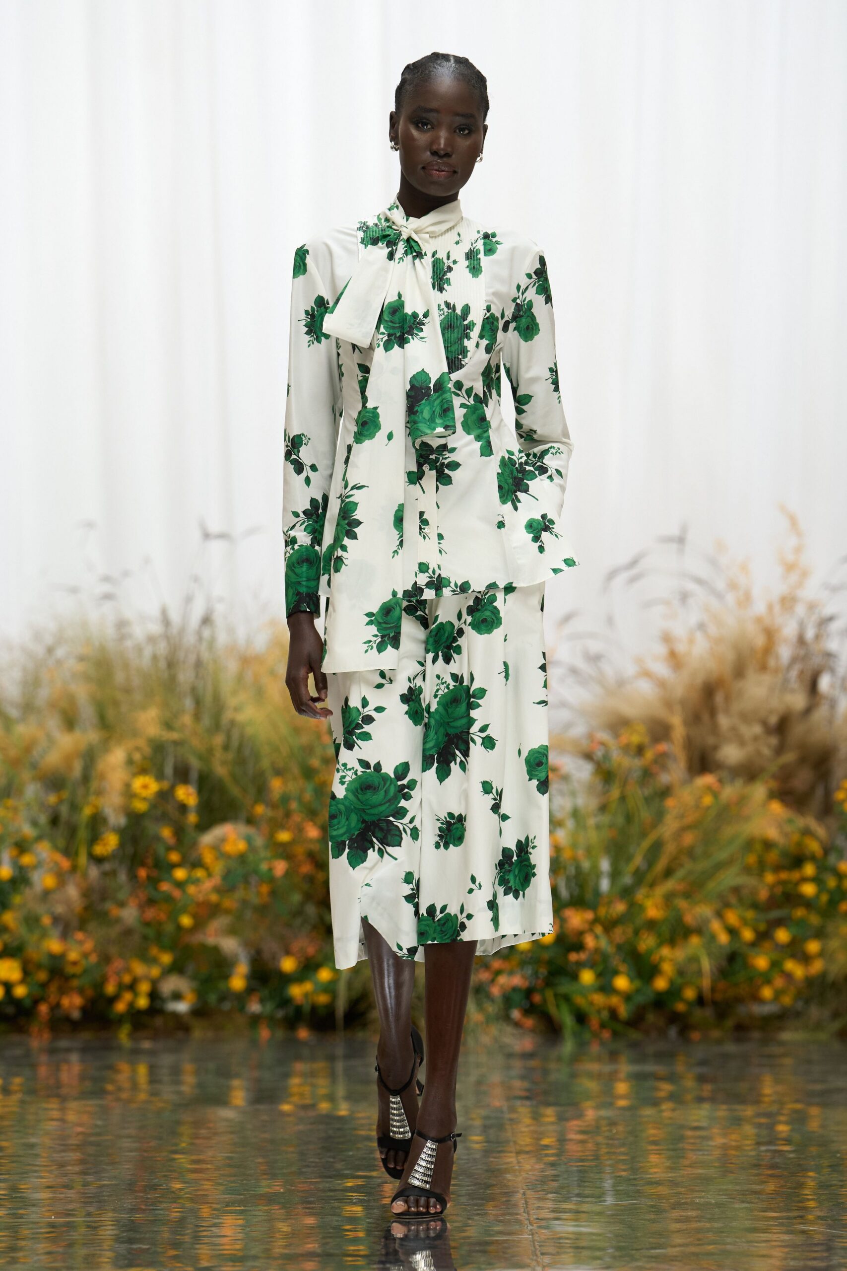 Ivory satin blouse scattered with emerald roses, long scarf tie draped loose. Matching bias-cut midi skirt, one uneven panel peeking below the hem. Black ankle-strap sandals with a ribbed silver column heel. Crossing the conservatory footbridge before the wedding guests find their seats.