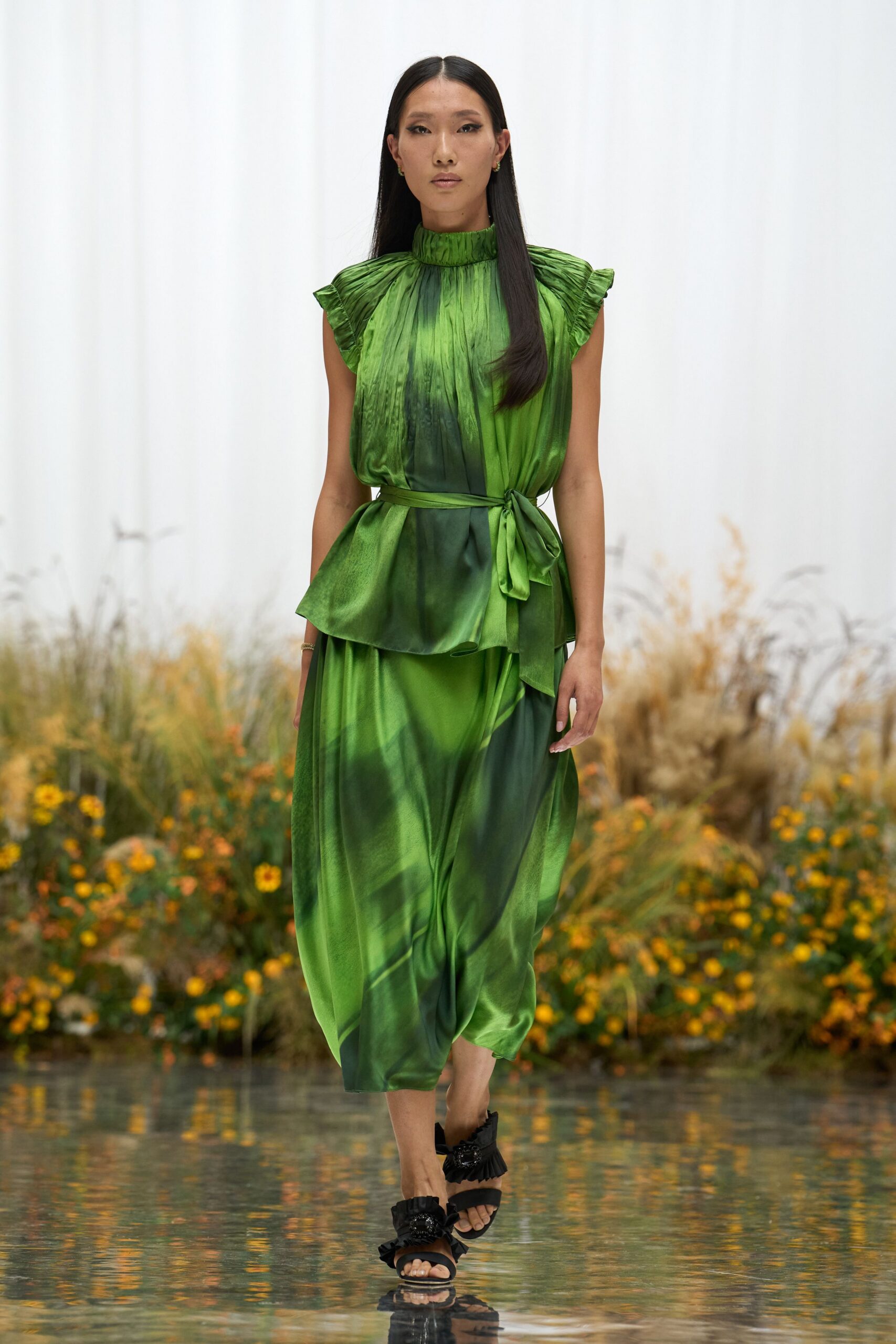 Leaf-green satin dress, ruched mock neck and flutter sleeves above a tied peplum waist. Black sandals with ruffled straps and faceted clusters at the vamp. Crossing the conservatory path after they have watered everything down.