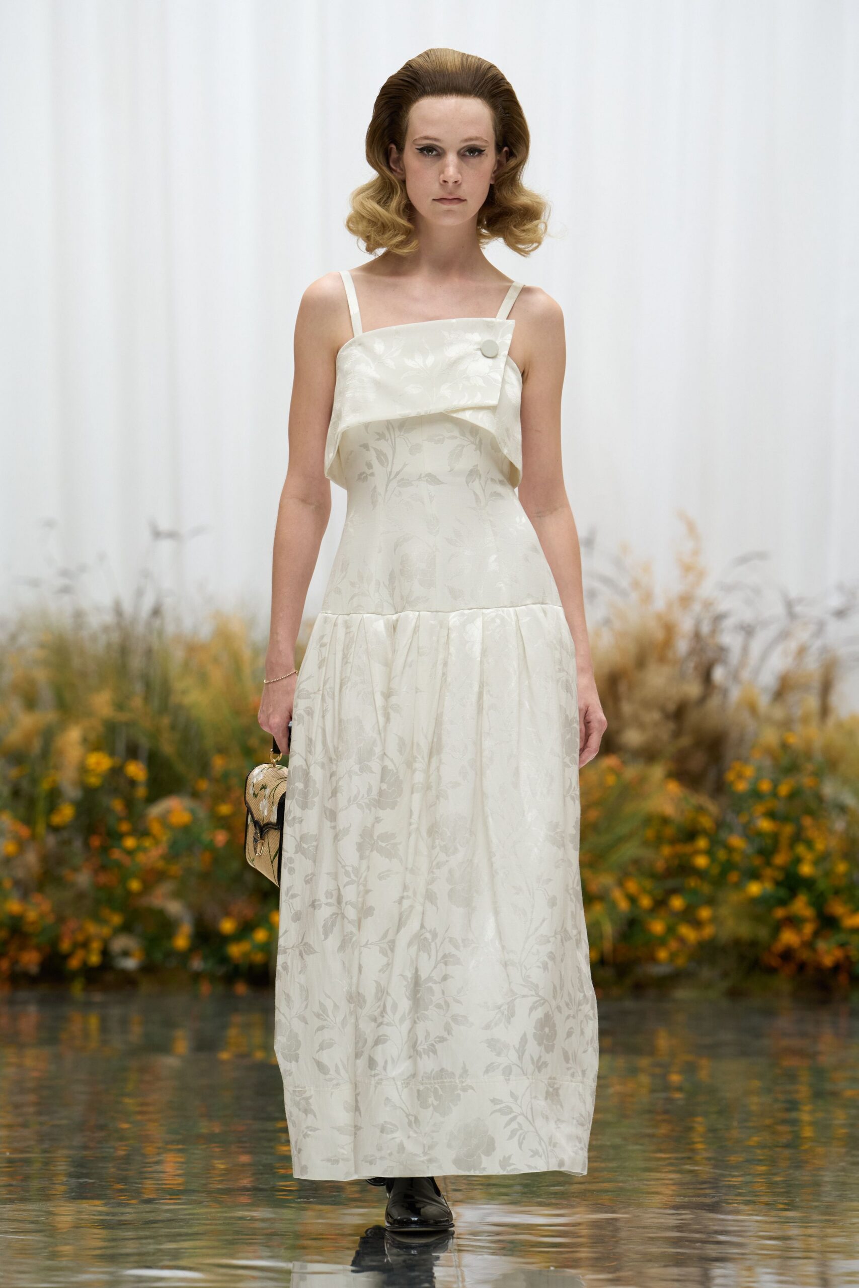 Ivory floral jacquard slip dress with a folded bib panel, one covered button. Fine gold chain bracelet. Mini tapestry box bag with black piping. Black patent square-toe shoes. Crossing the conservatory footpath after somebody else's afternoon wedding.