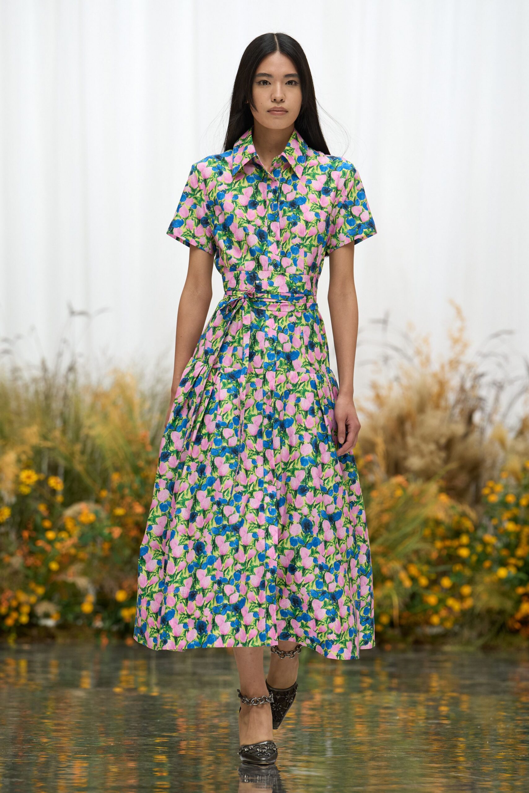 Pink and cobalt floral shirt dress, buttoned through with a self-tie waist knot. Black pointed pumps ringed with chunky silver chain at ankle and vamp. Crossing the conservatory path just after the watering hose was dragged through.