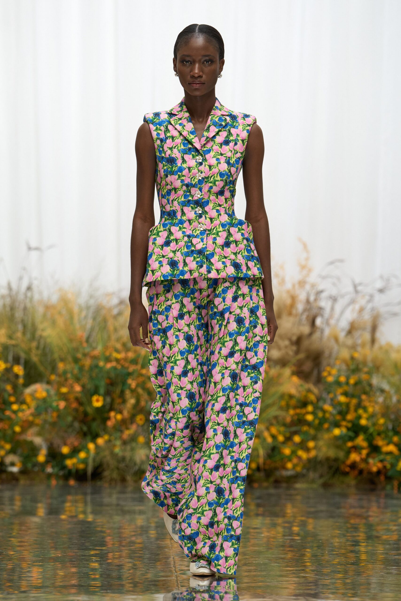 Tailored floral brocade vest, peplum hips jutting from the waist. Wide floral trousers in the same jacquard, pooling over the shoe. Small silver hoops. White pointed flats with jeweled bows. Crossing a conservatory footbridge while the koi disturb the water.