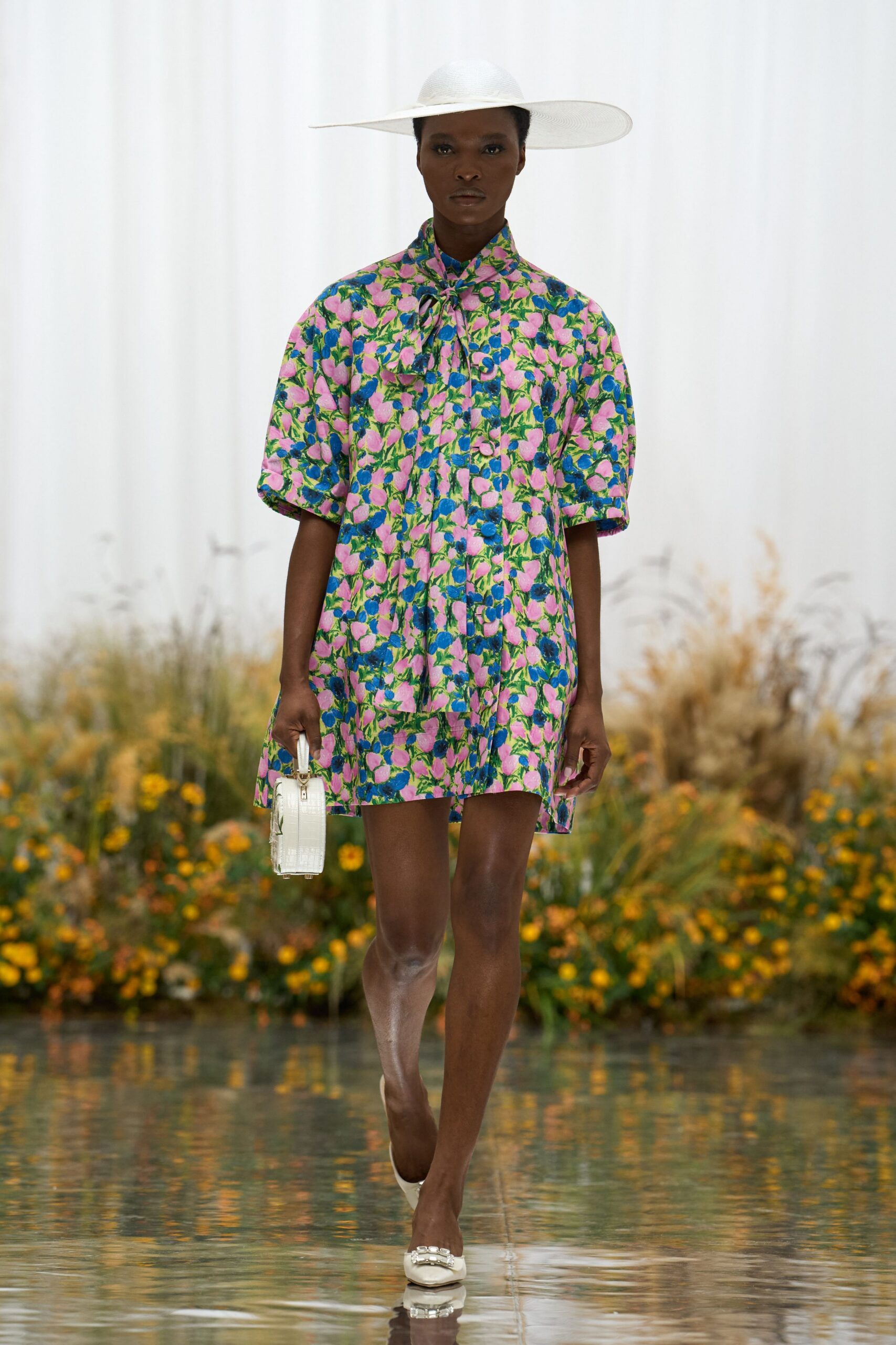 White woven sun hat with a brim stretched long to one side. Pink and cobalt floral jacquard shirt dress, tied neatly at the throat. White crocodile-embossed box bag with a short top handle. White pointed flats topped with a square crystal buckle. Walking beside a reflecting pool after the garden luncheon thins out.