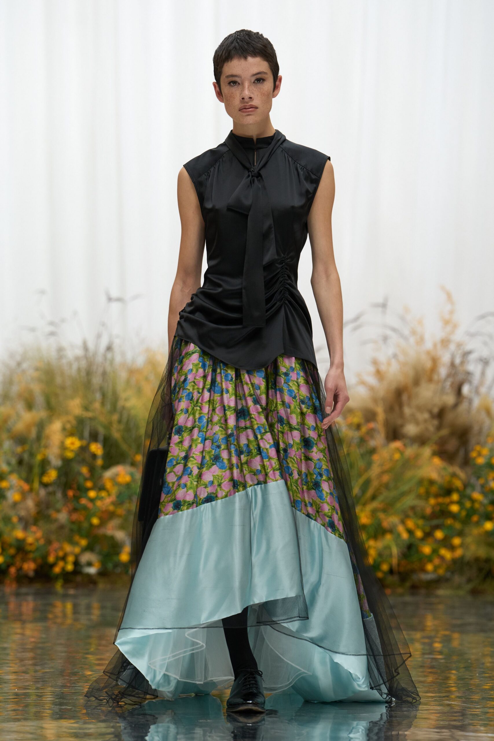 Black satin blouse with a narrow neck tie and ruched side seams. Floral satin skirt panelled with a pale blue hem under black tulle. Opaque black tights. Black leather lace-ups, square at the toe. Crossing the conservatory path after the watering cans have already been put away.