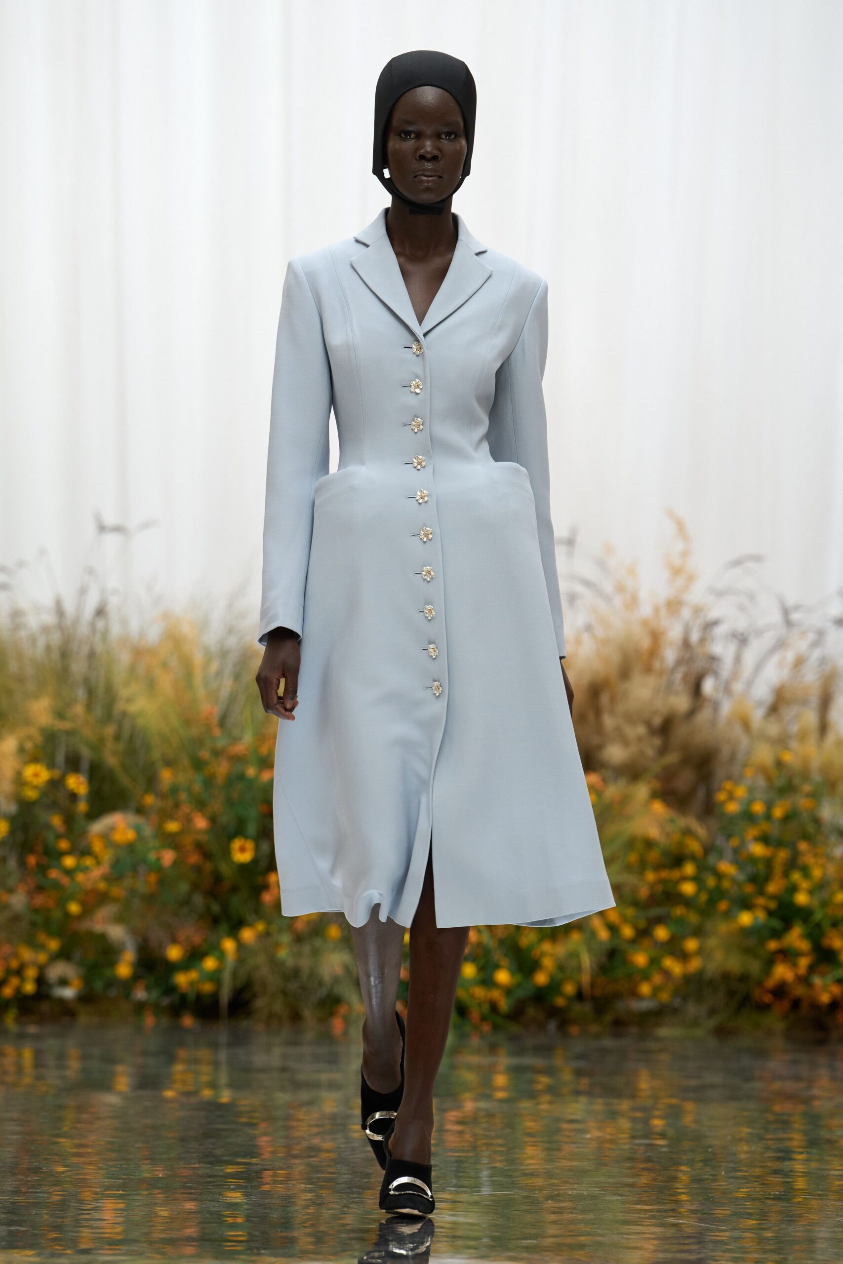 Black stretch hood fitted close under the chin. Powder-blue tailored coatdress with flower-shaped jeweled buttons and rounded hip pockets. Black pumps capped with curved silver bars. Crossing a glassy conservatory path after the gardeners have watered everything.
