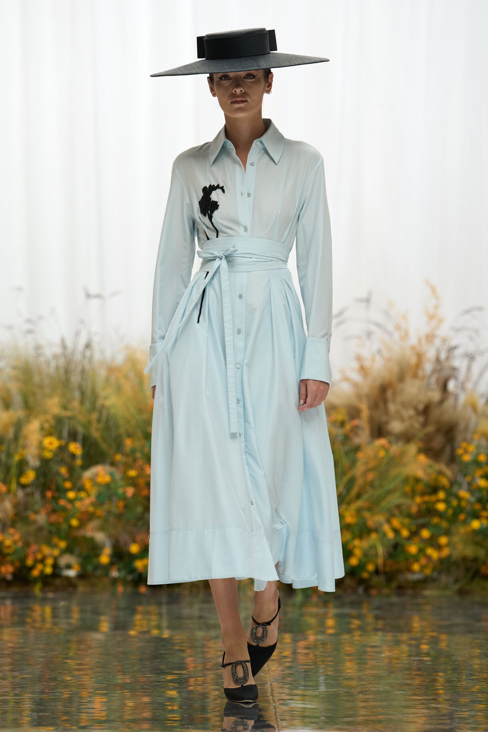 Flat charcoal boater with an extra-wide brim and tall grosgrain band. Pale blue poplin shirt dress, wrapped waist tie and black floral applique. Black Mary Jane pumps with oval buckles crusted in dark stones. Crossing the orangery path just after the gardeners have watered everything.