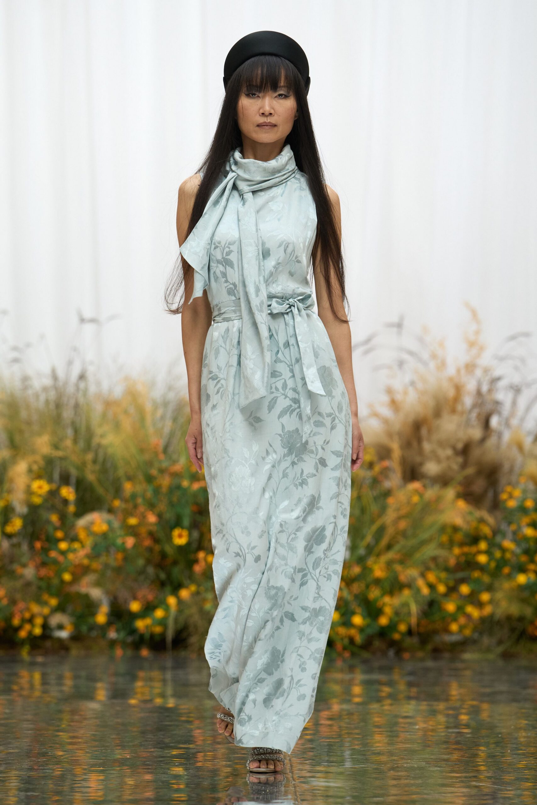 Matte black pillbox hat, rounded close over the crown. Pale blue floral jacquard jumpsuit with scarf neck and self-tie waist. Beaded flat sandals, narrow metallic straps at the toes. Crossing a shallow reflecting pool after the garden wedding guests have drifted off.