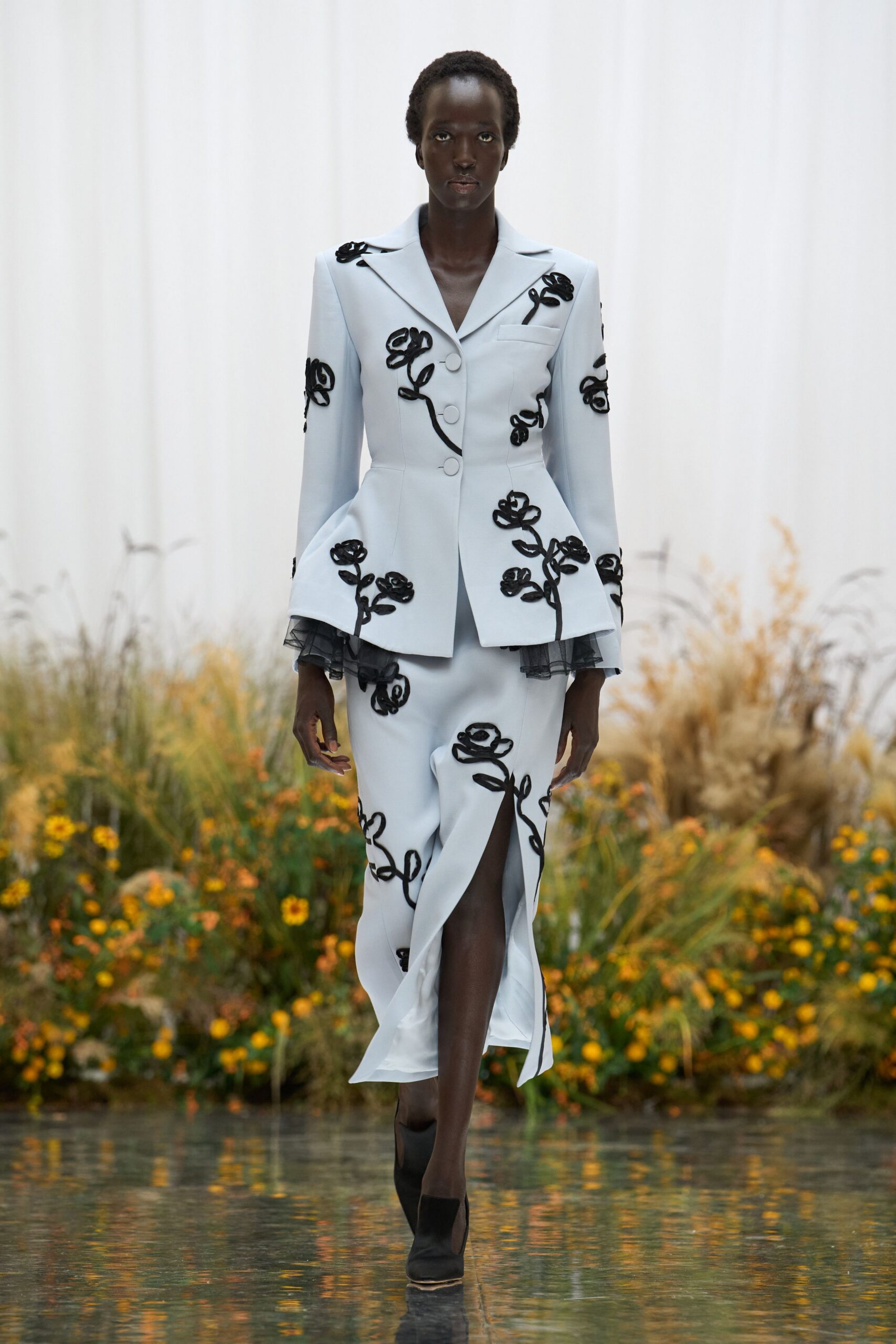 Powder-blue tailored blazer traced with black cord roses, tulle peeking at the cuffs. Matching narrow skirt split high at the front, the same corded blooms climbing. Black ankle boots with a pale sole edge showing. Crossing the conservatory floor just after the watering hoses were dragged away.