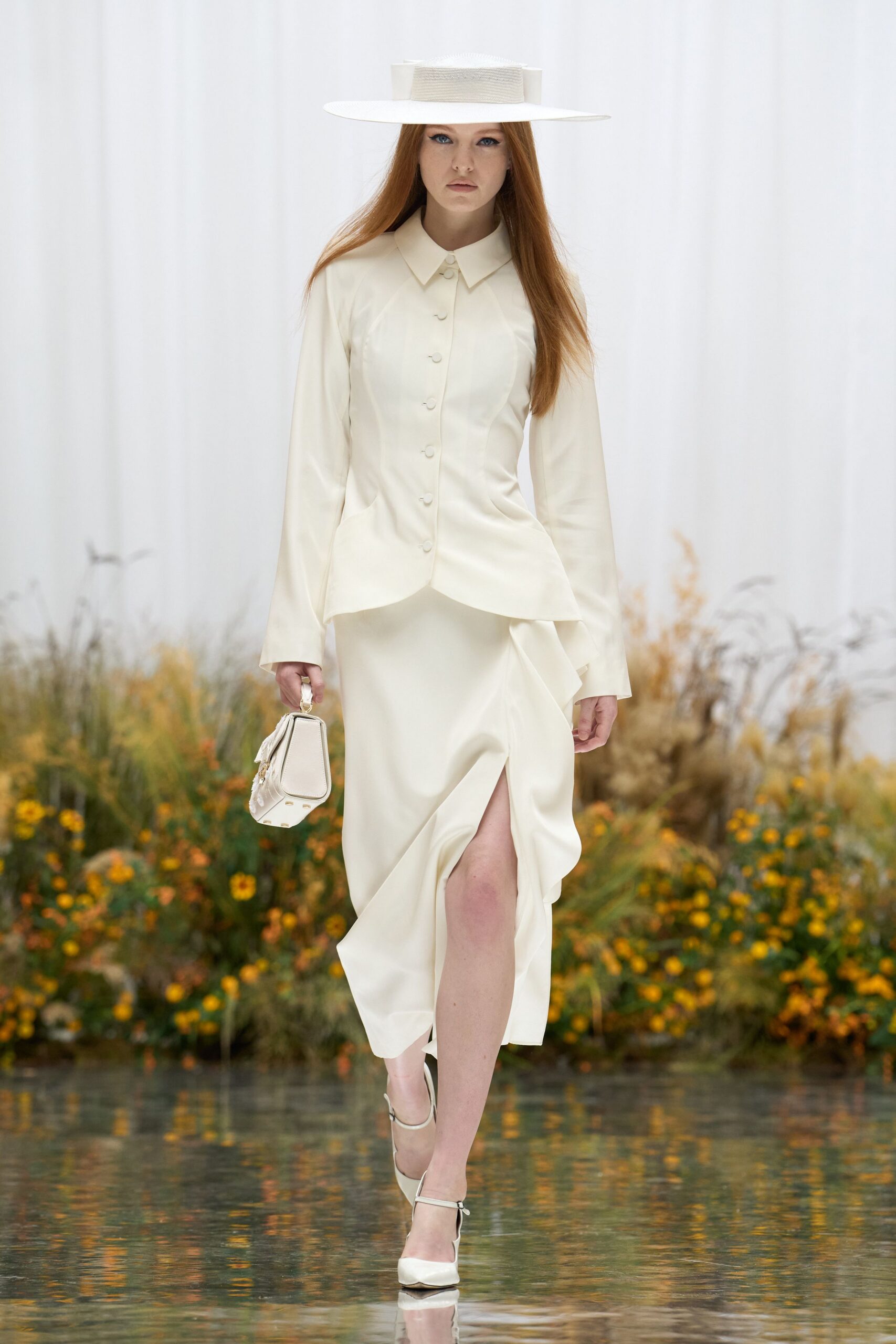 Flat-brim ivory boater with a grosgrain-wrapped crown. Cream sateen blouse, pointed collar and covered buttons to the hip. Bias-cut ivory skirt with a high slit and twisted drape. Small top-handle bag with a tasseled side seam. Ivory ankle-strap pumps. Crossing the orangery after the wedding guests have drifted outside.