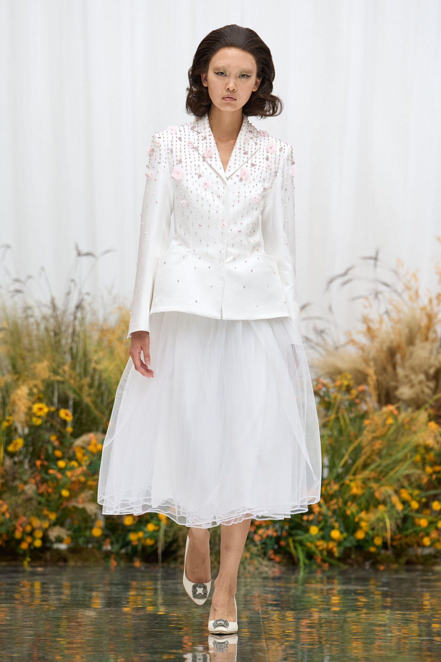 White satin jacket, shoulders dusted with pink rosettes and crystal beading. Calf-length white tulle skirt, layered mesh floating away from the hips. Ivory pumps with square crystal buckles. Garden wedding before noon, grass still wet through the hem.