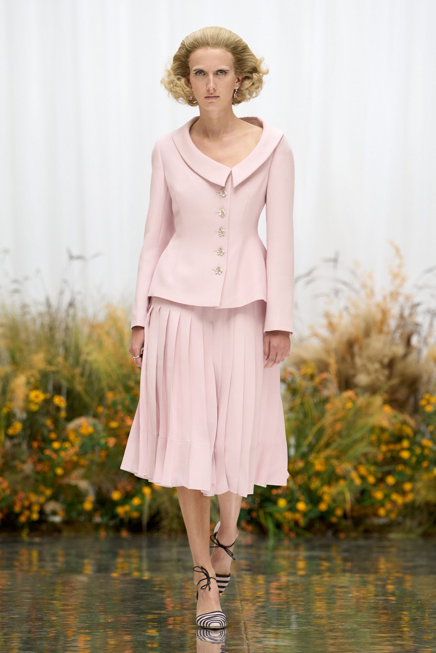 Powder-pink tailored jacket with a wide rolled collar and jeweled buttons. Matching midi skirt in soft knife pleats, hem wavering unevenly. Black-and-white striped pumps laced over the instep. Crossing a conservatory aisle while the orchids are being watered.