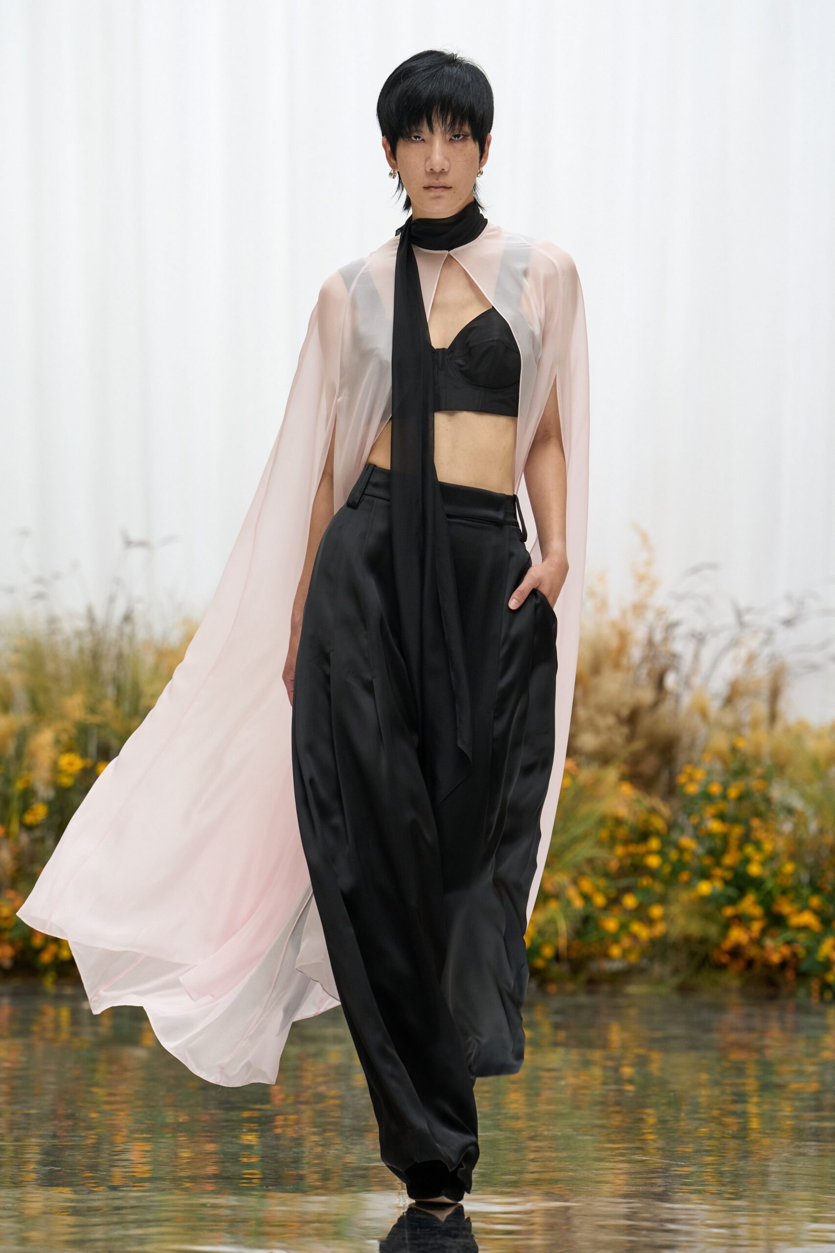 Blush sheer chiffon cape top, slit to hang in long winged panels. Black georgette scarf knotted close, one tail dropped past the waist. Black bralette with sculpted cups and a narrow center seam. Black satin trousers cut wide and full through the pleated front. Crossing a garden path after rain, hem picking up the pond light.