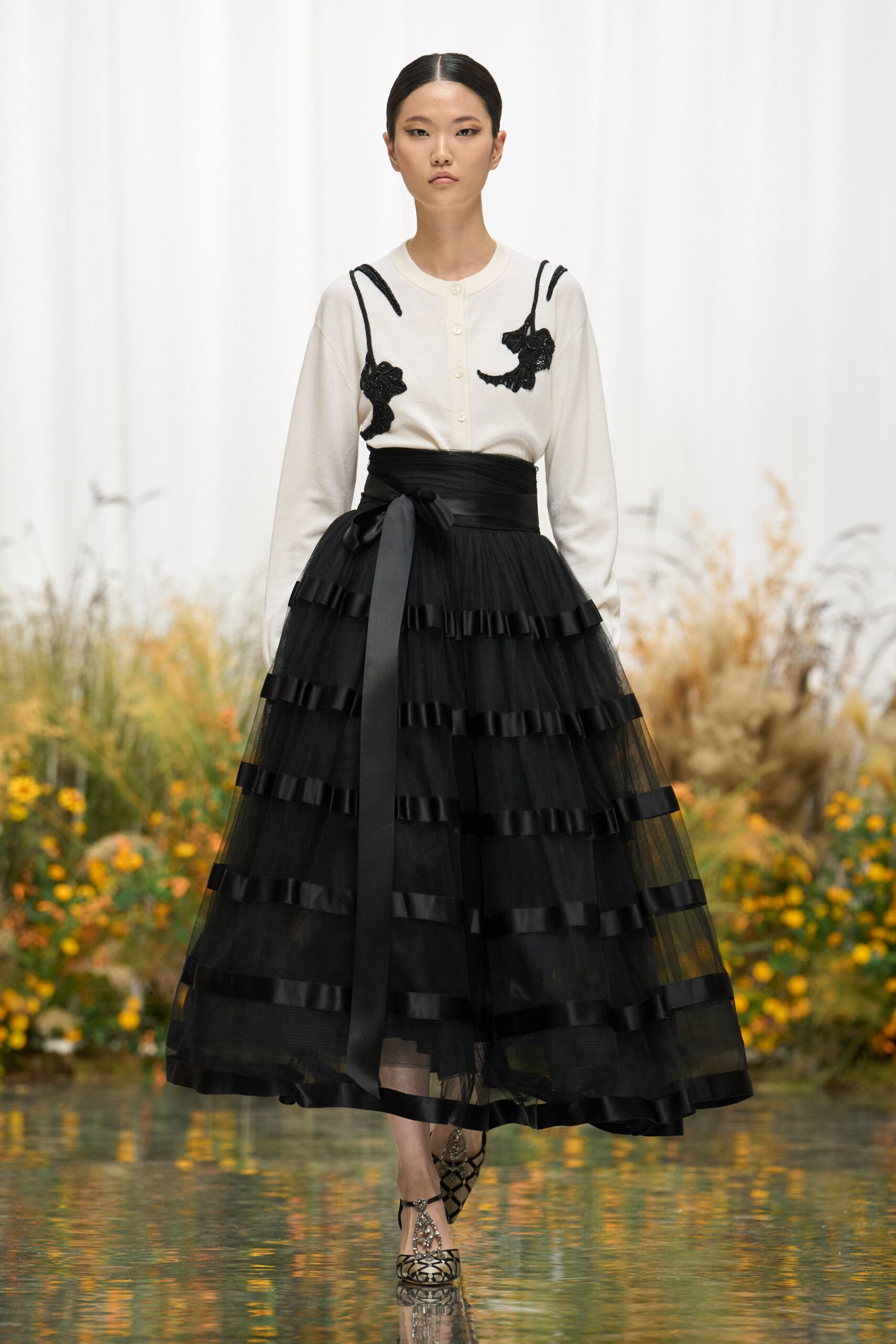 Ivory fine-knit cardigan with black beaded floral cords curling off the shoulders. Black tulle midi skirt banded in satin ribbon tiers, sash tied low at the waist. Black lattice heels with crystal loops climbing the ankle. Crossing the orangery after rain, trying not to catch the hem.