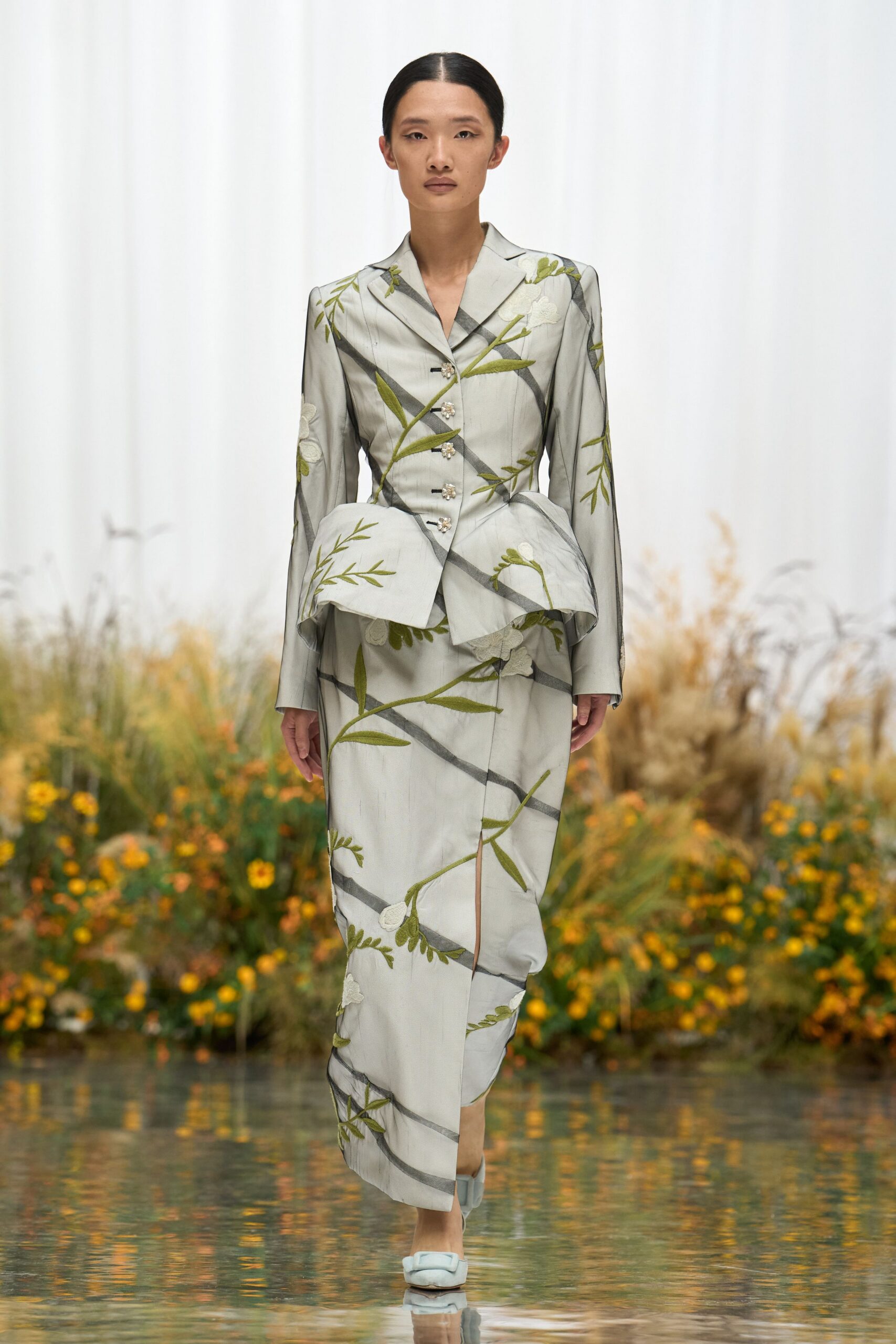 Pearl-grey tailored jacket with olive vine embroidery and a flared peplum. Matching column skirt, front slit breaking the diagonal stem pattern. Pale satin mules with squared, padded bows. Crossing a glassy courtyard after rain, flowers blown against the walls.