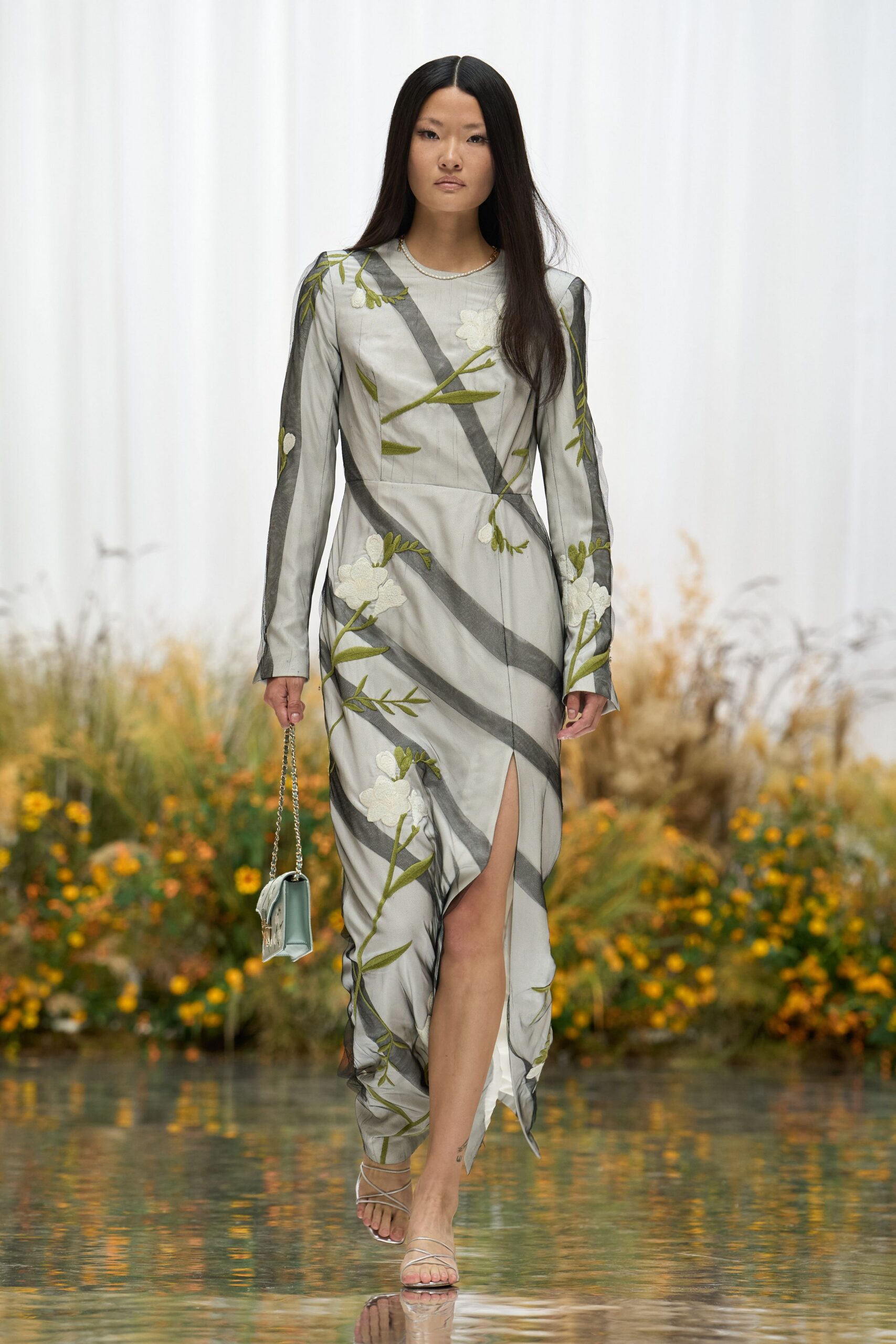 Pale grey long-sleeved dress with raised white floral embroidery and smoky diagonal bands. Icy box bag on a fine silver chain. Barely there silver straps winding across the toes. Crossing a conservatory footbridge while the pond water catches the light.