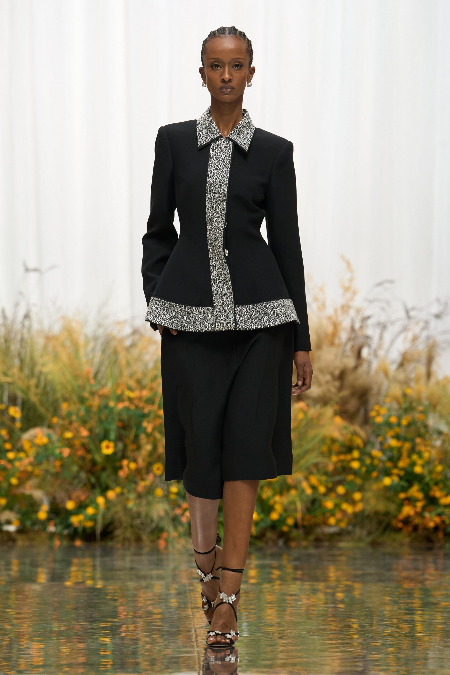 Small silver hoops at each ear. Black fitted jacket with crystal-packed collar, placket and curved hem band. Black culottes cut mid-calf with a soft front crease. Black lace-up sandals dotted with crystal flower clusters. Crossing the conservatory path after dark, hem skimming reflected water.