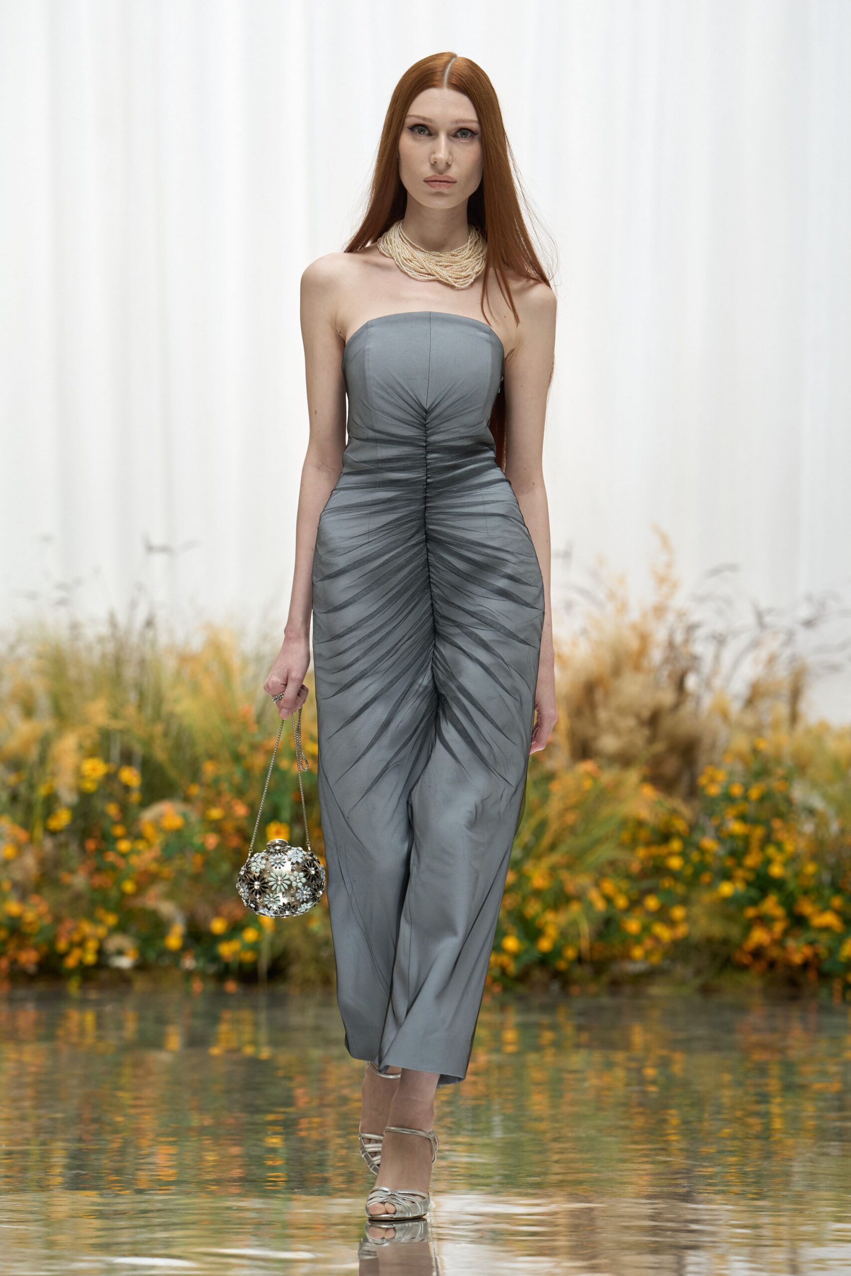 Dove-grey jumpsuit with a gathered center seam and smoky leaf-like shading. Multiple cream bead strands pooled close around the collarbone. Silver sphere minaudiere crusted with small flower-like studs. Pale metallic straps crossing the toes. Crossing a glassy garden pond while the last asters are still out.