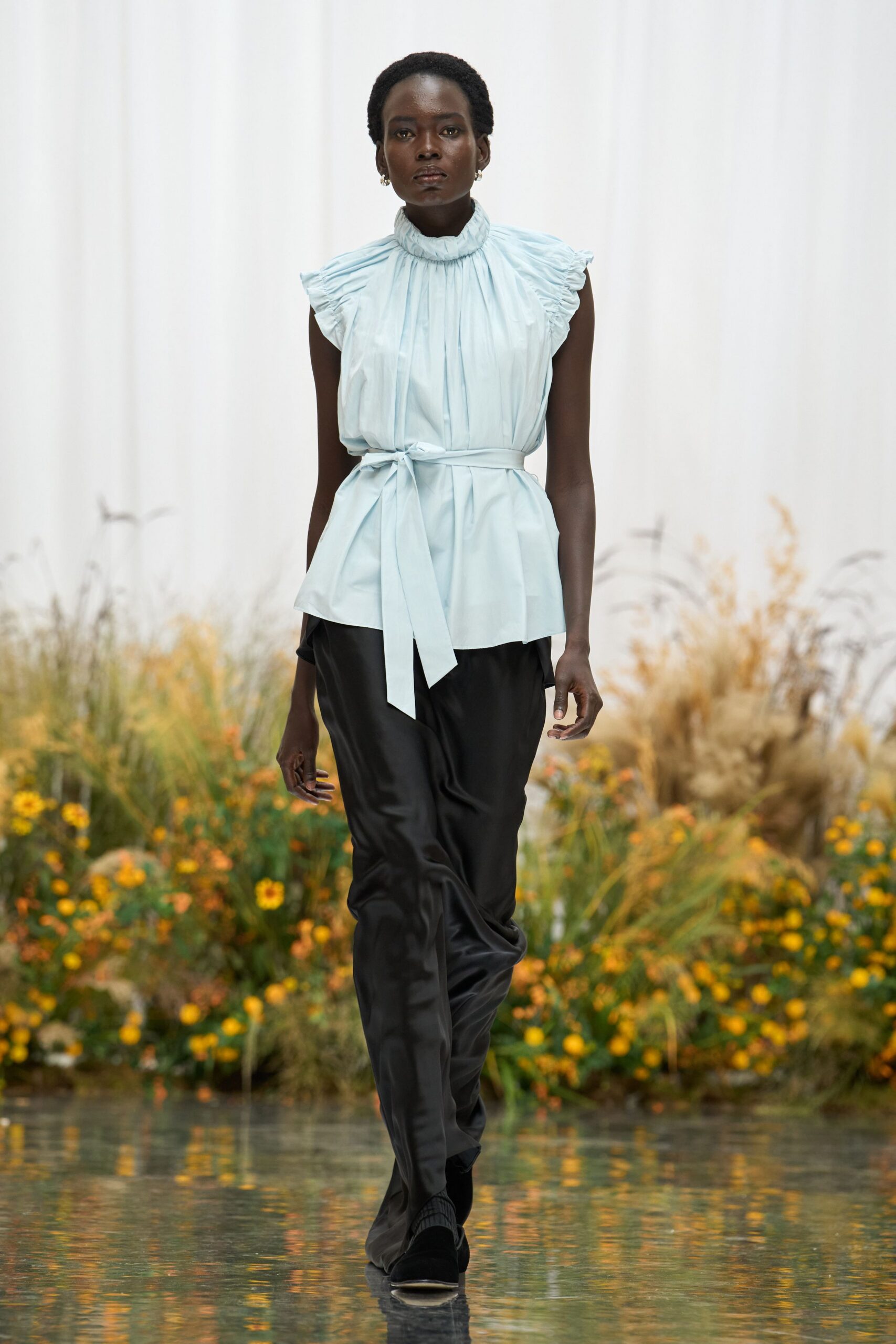 Pale ice-blue blouse with a ruffled stand collar and flutter sleeves. Self-tie sash knotted off-center, one end hanging long. Black satin trousers falling in a loose, liquid drape. Black boots with ribbed knit shafts peeking under the hem. Tiny silver hoops. Crossing the conservatory path before the sprinklers start again.
