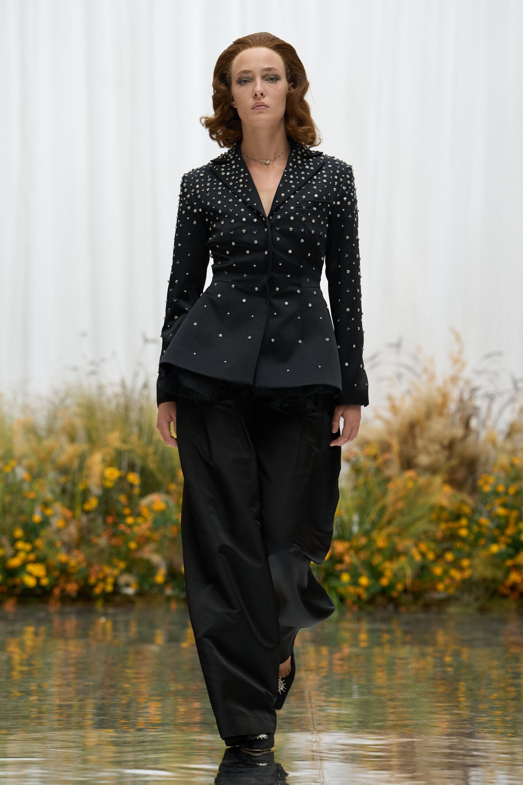 Fine silver chain with a tiny irregular drop at the throat. Black tailored jacket strewn with crystal studs, peplum kicking out sharply. Loose black satin trousers, pooling and catching light in soft folds. Black slip-ons with a starburst brooch at the vamp. Crossing a conservatory path after rain, gravel still stuck to the hem.