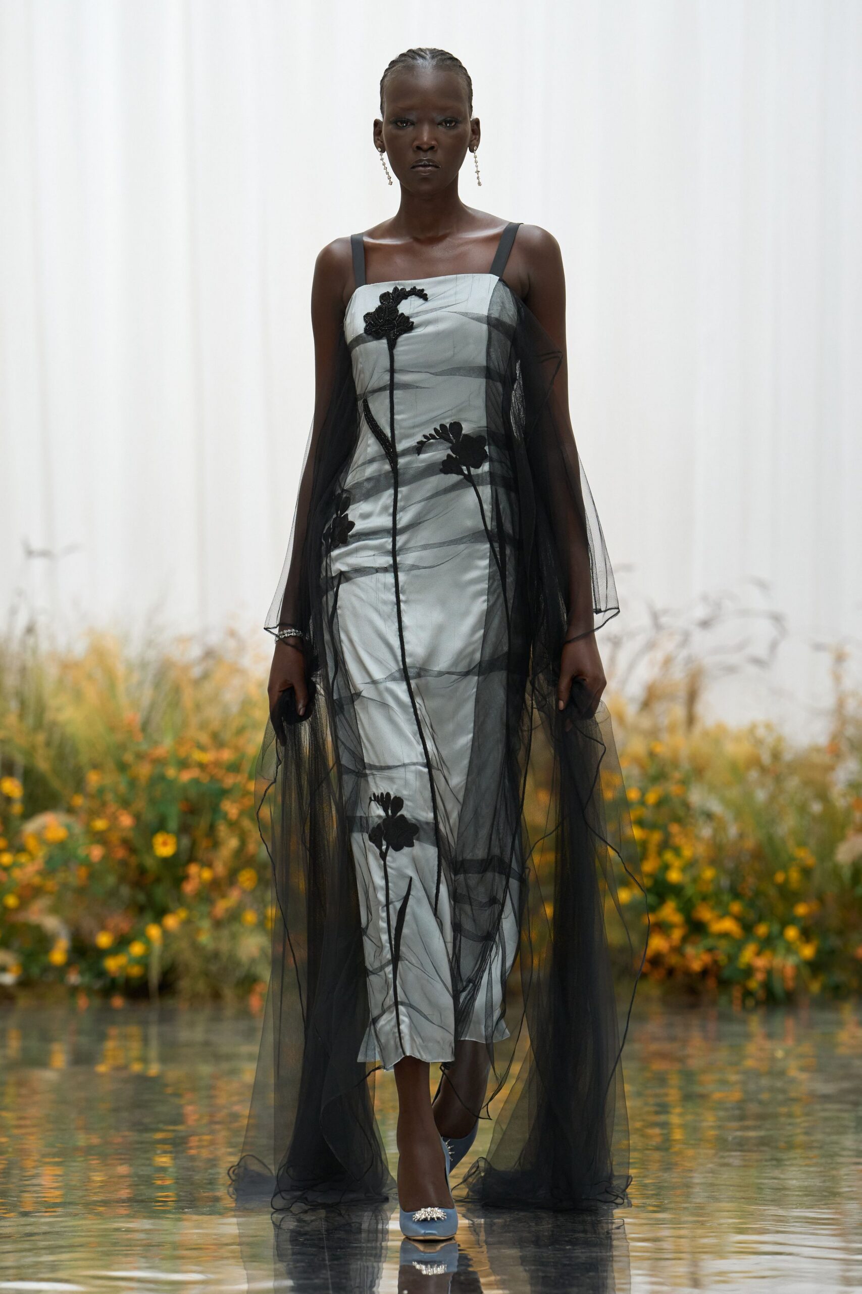 Pale silver satin column dress with black floral appliqué and wiry stems. Sheer black tulle falling from the straps in long uneven panels. Fine drop earrings. Powder-blue pumps with a crystal burst at the toe. Crossing the conservatory pond while the last marigolds are still up.