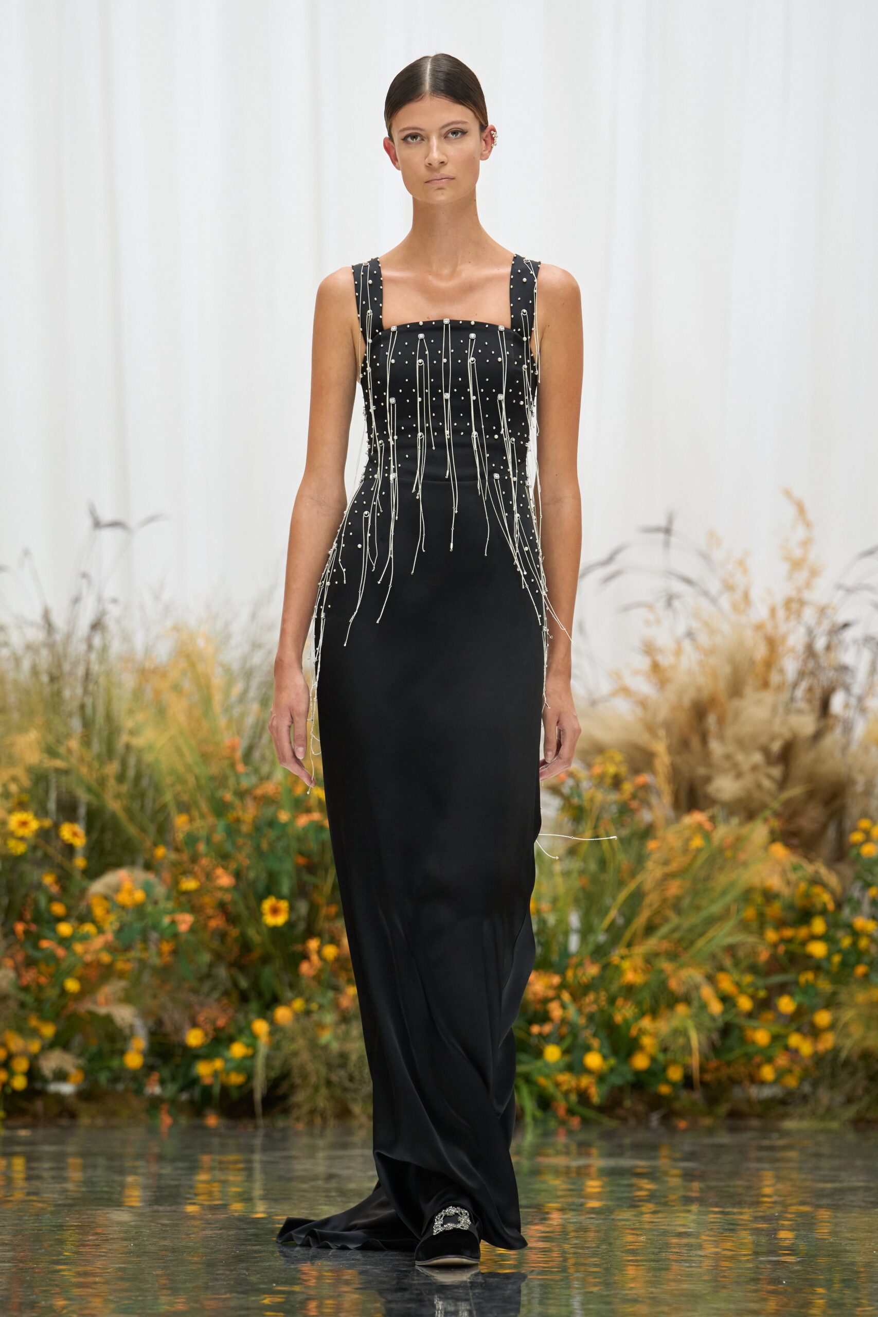 Black floor-length column dress with silver beadwork and dangling cord strands. Black velvet slippers topped with a clustered crystal buckle. Crossing a conservatory path just after the watering hoses were dragged through.