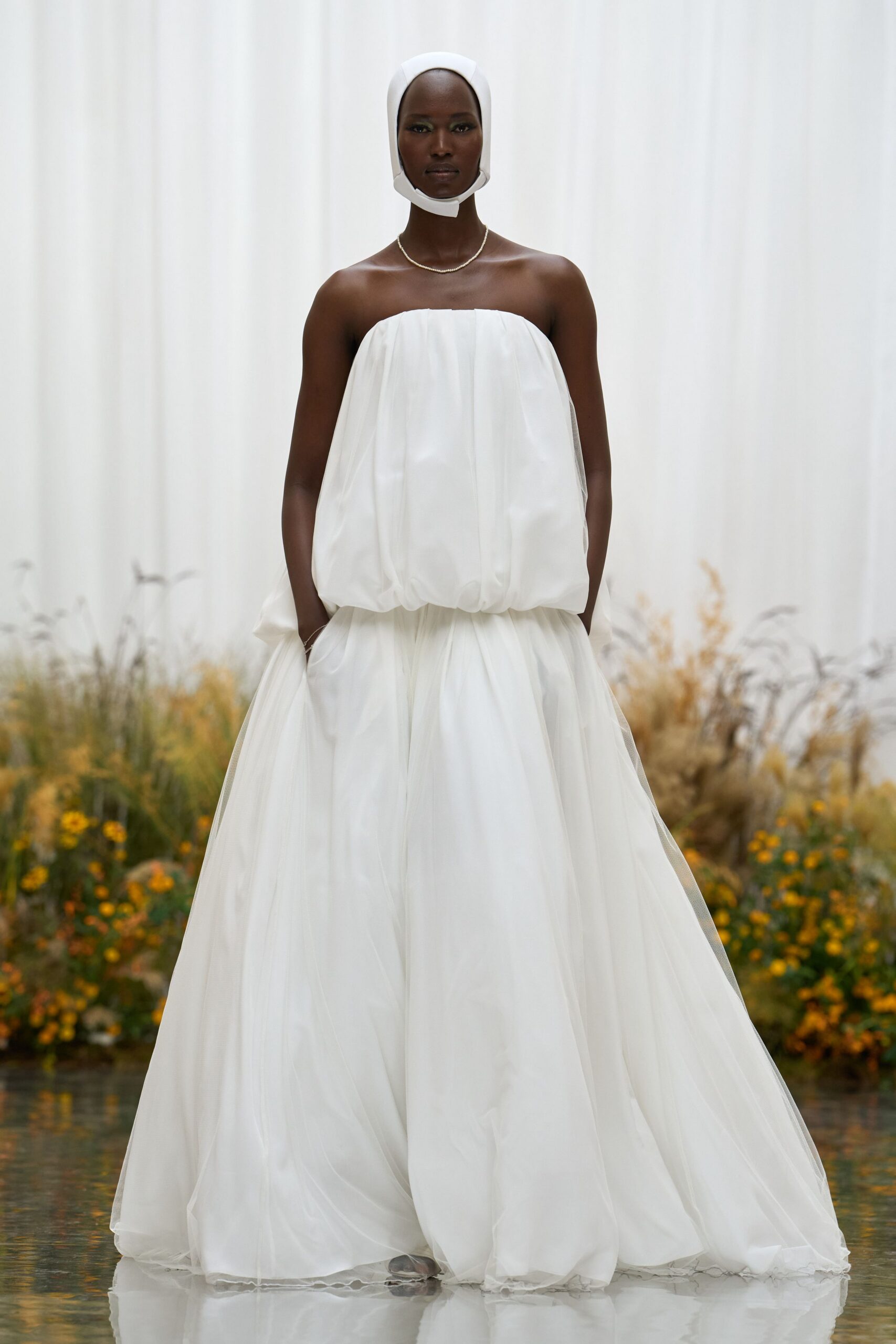 White padded bonnet framing the jaw with a cutaway chin band. Short rope of small pearls. Strapless ivory gown in airy tulle, bubbled at the waist and pooling wide. Crossing a conservatory aisle while the gardeners are still watering.
