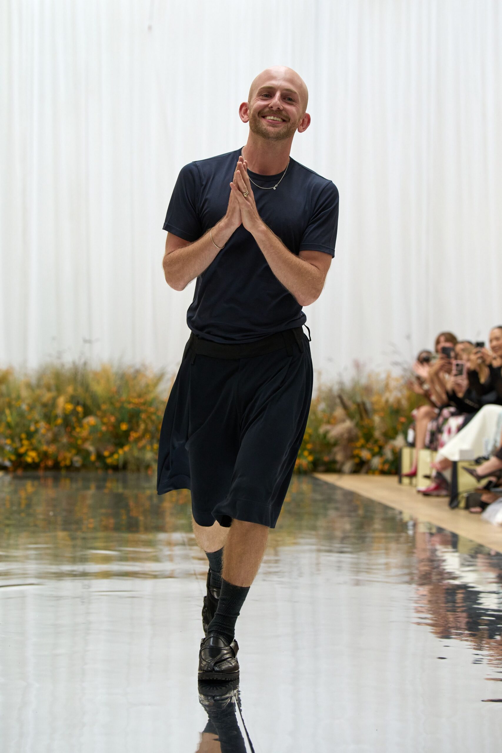 Dark navy jersey tee with a close crewneck and clean short sleeves. Voluminous black shorts with deep pleats falling from a wide waistband. Black ribbed mid-calf socks. Black leather penny loafers with a heavy lug sole. Walking the edge of a shallow reflecting pool after the speeches end.
