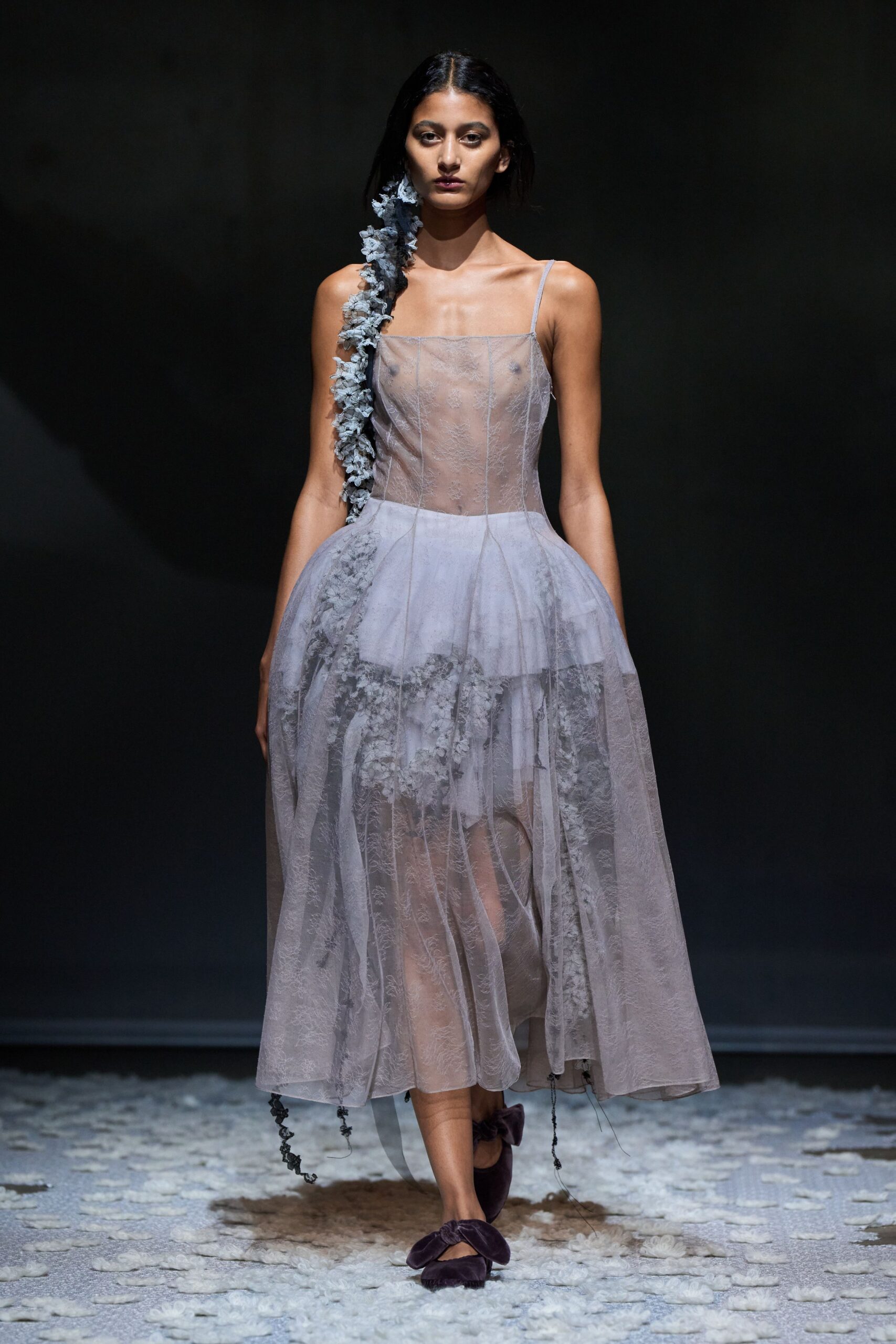 Dusty lilac organza dress with sheer boned bodice and floating embroidered underlayer. Grey fabric blossoms trailing from ear to hem, ending in loose cords. Plum velvet flats with oversized bow knots. Walking across a chapel aisle after the flowers have already dropped.