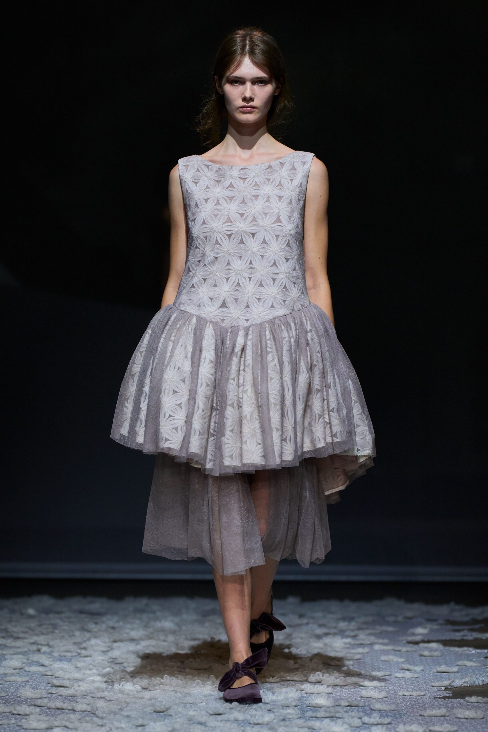 Pale grey jacquard dress with petaled starburst motifs and a dropped waist. Sheer tulle pannier overskirt, puffed at the hips and falling unevenly. Dusty mauve flats with oversized velvet bows across the instep. Crossing a winter garden after the last wedding guest has gone.