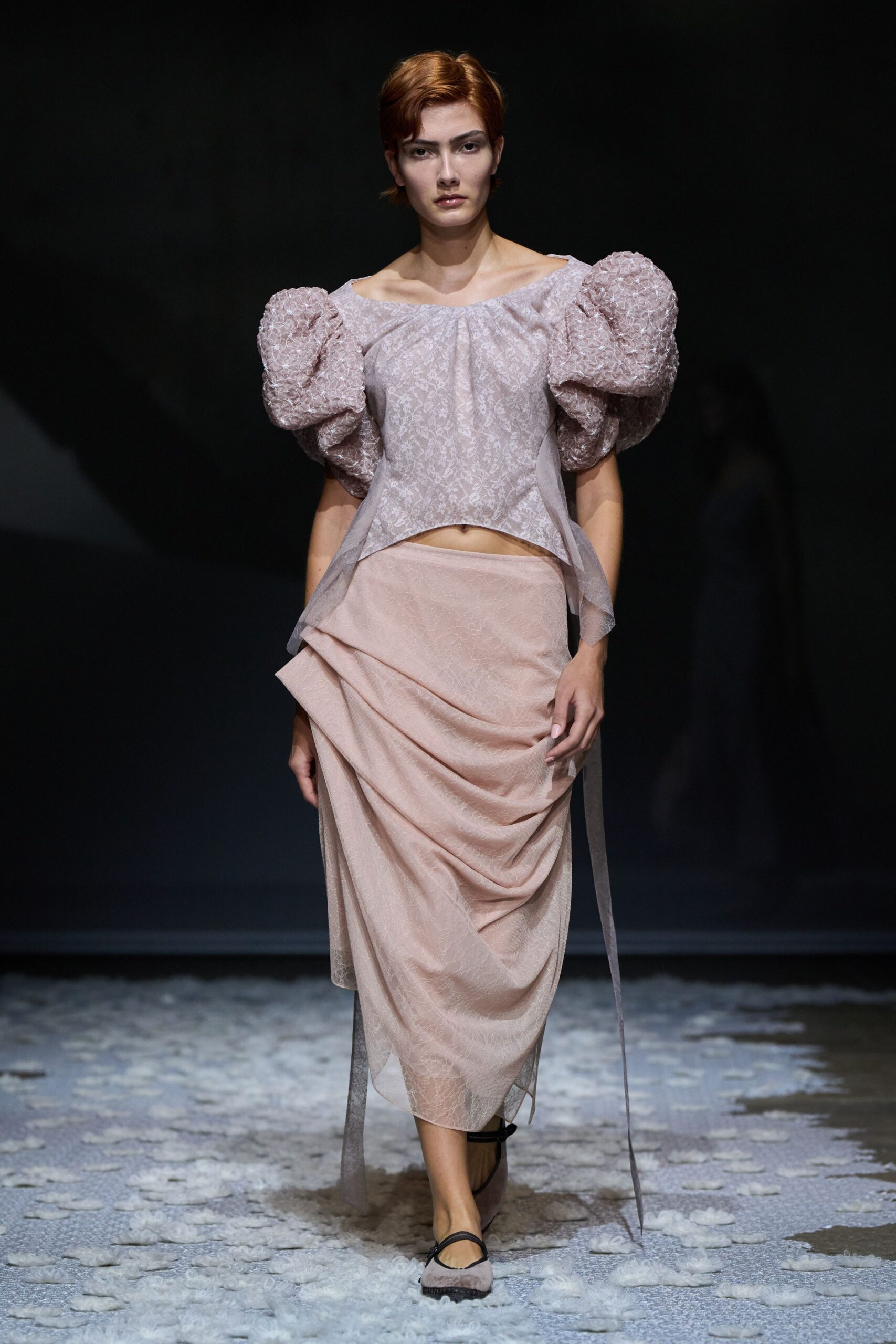 Blush floral jacquard blouse with oversized crumpled puff sleeves. Matching draped midi skirt in sheer layered organza, tied panel trailing. Pale mesh flats with thin black crossing straps. Walking across a wedding hall after the chairs have been stacked.