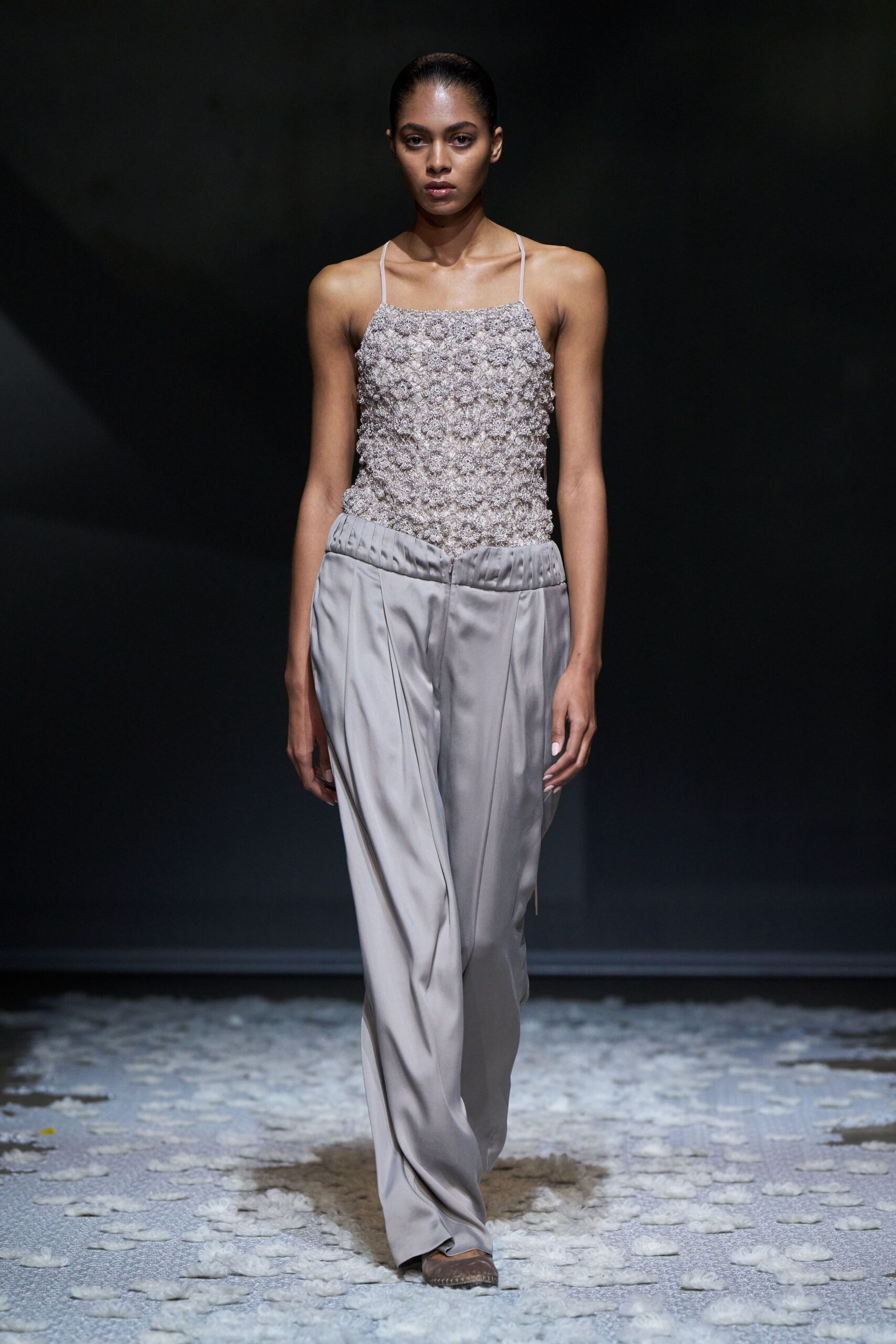 Pale beaded camisole densely crusted with rosette knots, held by fine straps. Pearl-grey satin trousers with a padded waistband and deep front pleats. Pointed suede flats peeking from the pooling hem. Walking across a ballroom floor after the confetti has gone damp.