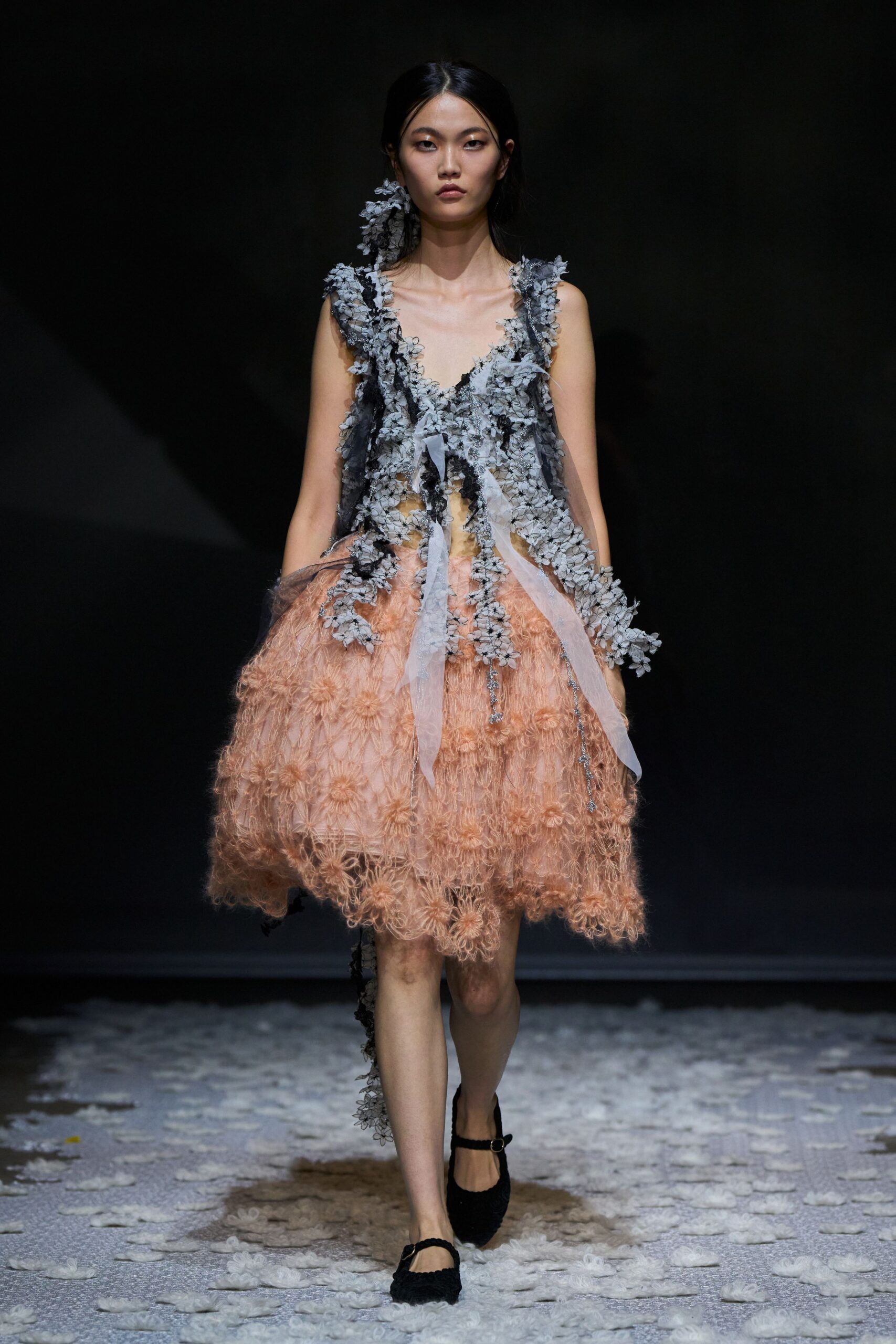 Blush cocoon dress with looped floral crochet swelling out at the hem. Grey petal-appliqué tulle draped in ragged garlands, organza ribbons hanging loose. Matching blossom cluster climbing one ear. Black Mary Jane flats with a single slim strap. Crossing a courtyard after the wedding, petals stuck to the damp paving.