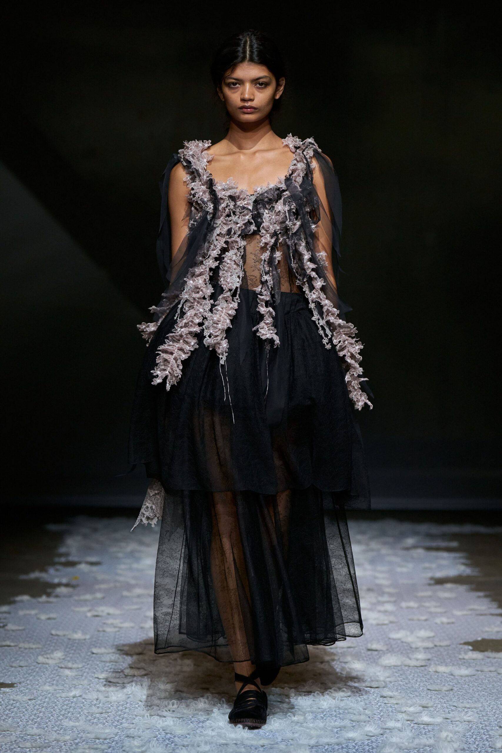 Black sheer tulle dress with mauve lace garlands trailing from the bodice. Black floral lace stockings visible through the transparent skirt. Black wrap flats with crisscross straps over the instep. Crossing a chapel aisle after the flowers have started to drop.