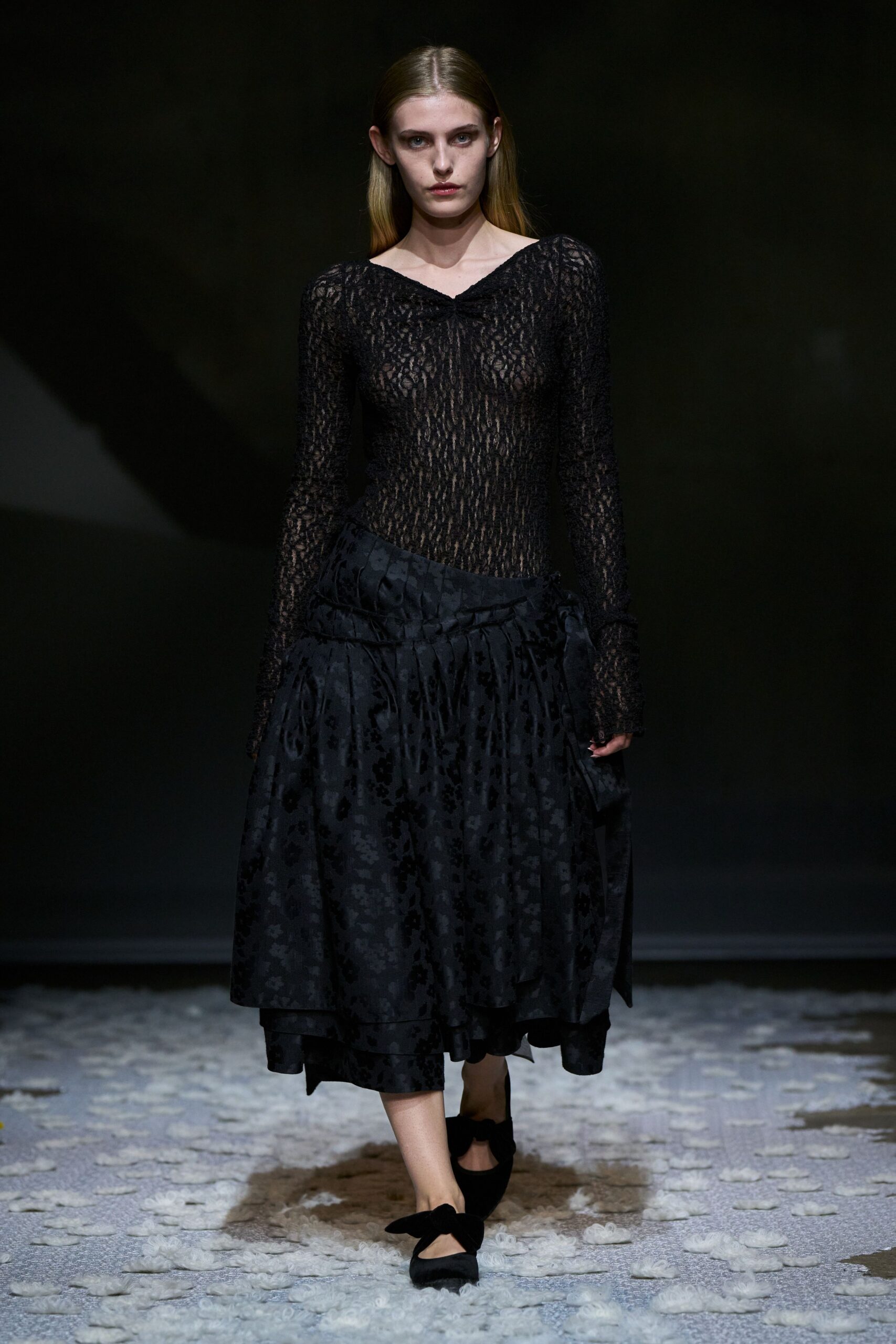 Sheer black openwork knit top, pinched once at the shallow V. Black jacquard skirt with dropped pleats and a wrapped overskirt panel. Black flats with oversized bow straps across the instep. Crossing a museum courtyard just after rain, hem skimming the wet stone.