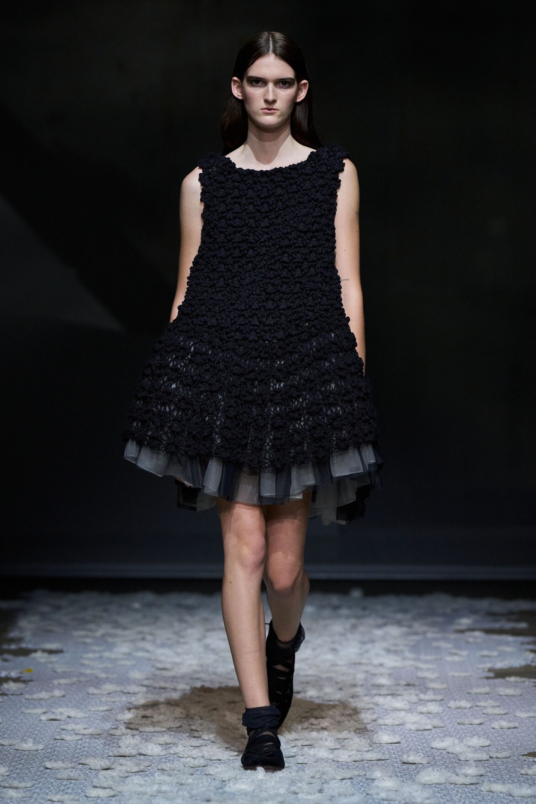 Black sleeveless dress crusted in nubby rosette clusters, grey tulle kicking from the hem. Black wrap flats with crossed straps climbing the ankle. Walking across a hall strewn with burnt paper snow after midnight.