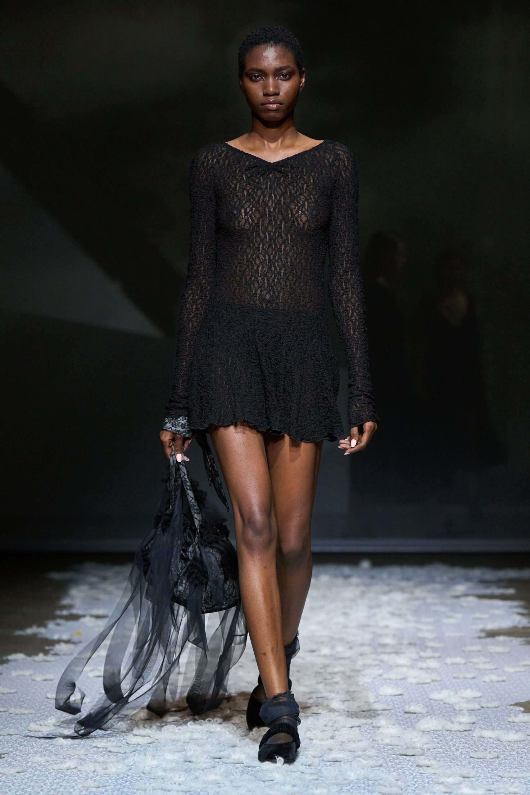 Black open-knit mini dress, sleeves pooled and hem left uneven. Black lace bag trailing long strips of smoky tulle. Black mesh flats with a wrapped strap across the instep. Crossing a hotel lobby barefoot in spirit, shoes still technically on.