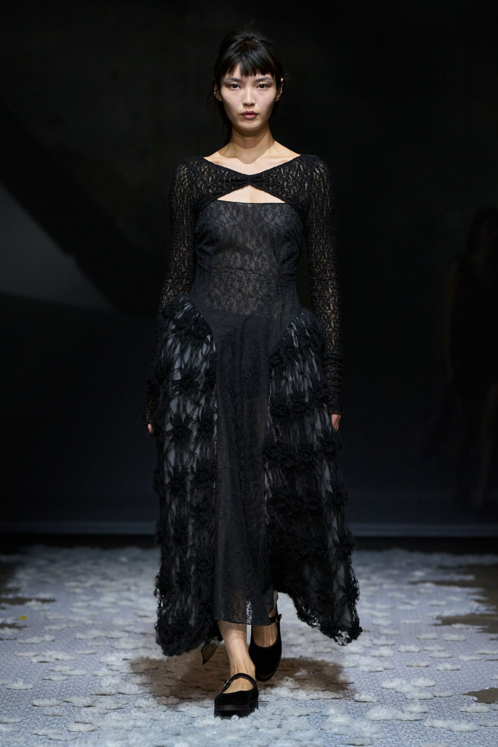 Black lace dress with a twisted cutout neckline and shaggy appliqued side panels. Black velvet Mary Janes with a thin strap across the instep. Crossing a hotel ballroom after the florist has swept up.