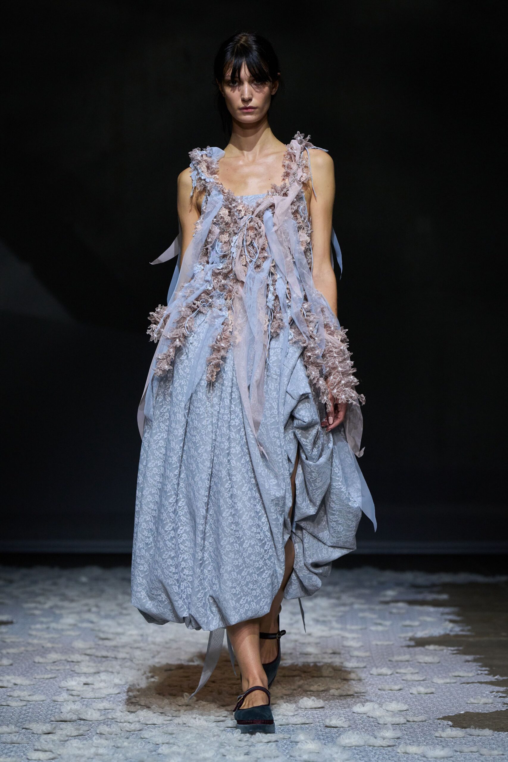 Powder-blue floral jacquard dress, skirt ballooned and hitched into a side swag. Blush tulle and frayed rosette mesh trailing in long loose ribbons. Dark mary jane flats with a thin instep strap. Walking across a hall after the wedding flowers have already been trampled.