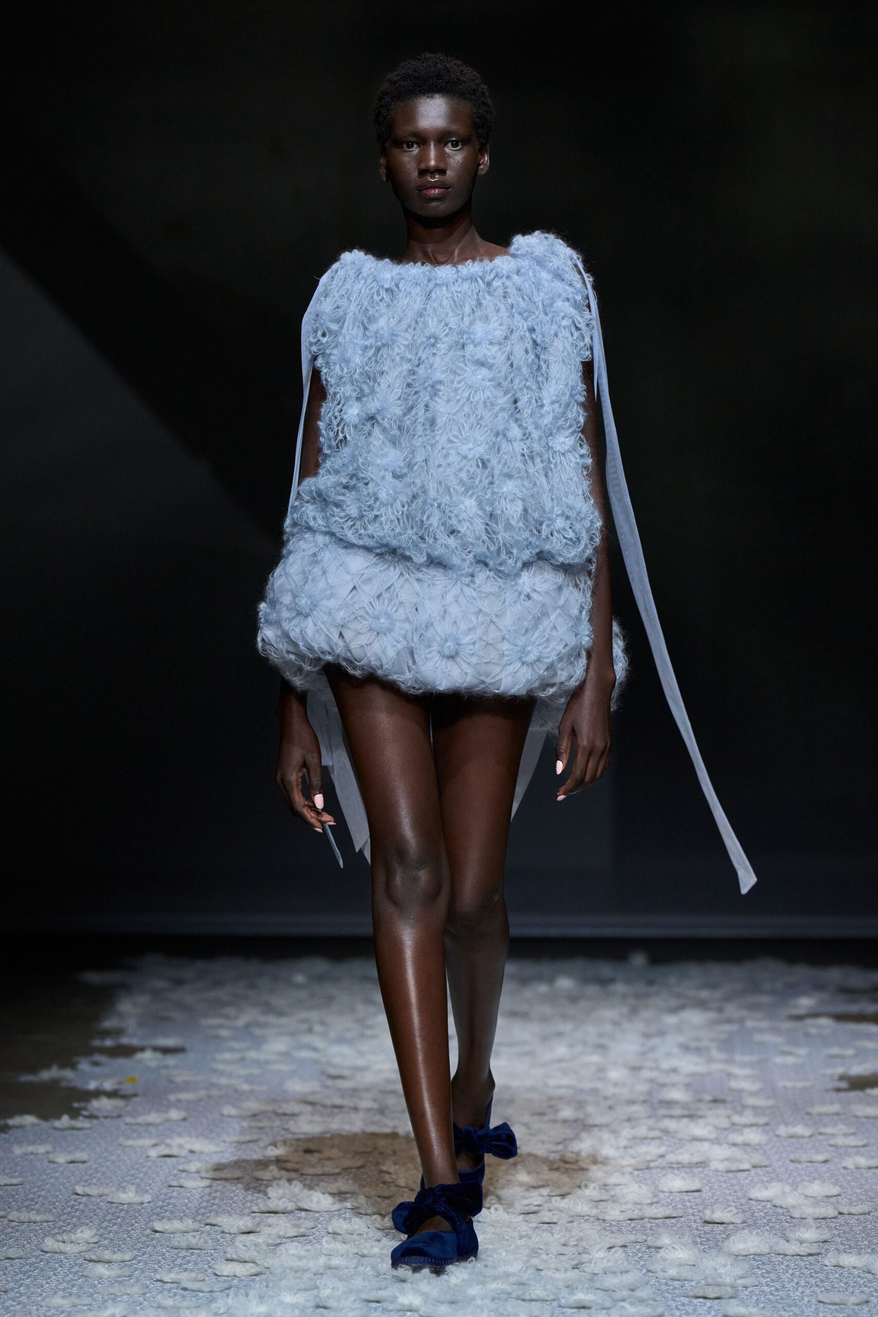 Powder-blue boucle clouding the bodice, with sheer ribbon trails from the shoulders. Bubble hem in smocked tulle, flower clusters pressed into the skirt. Cobalt velvet flats with crushed bows at the toes. First frost in the glasshouse, petals stuck damp to the path.