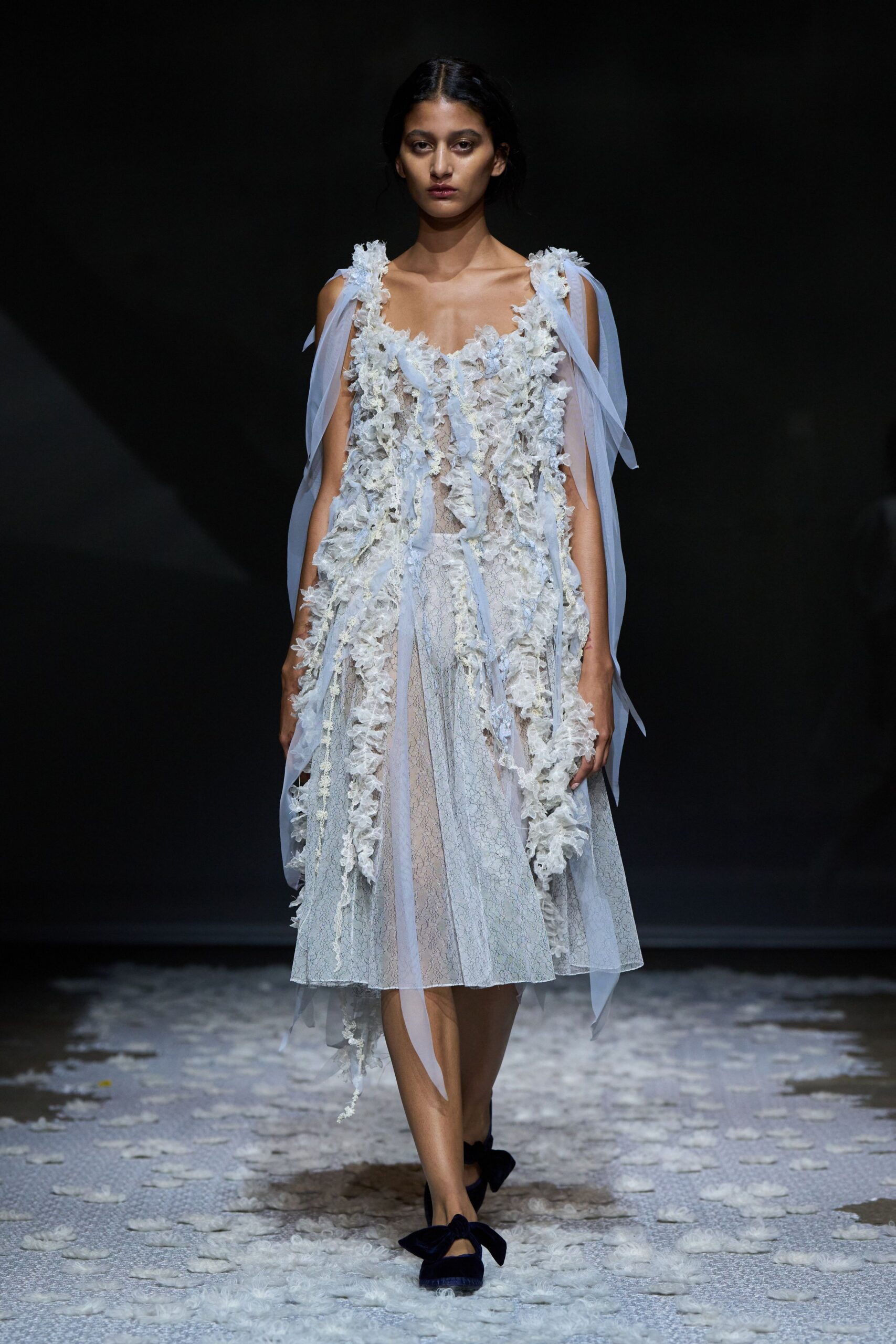 Pale lace dress veiled in scattered chiffon petals and long ribbon streamers. Black velvet flats with oversized bow knots at the toes. Crossing a chapel garden after the petals have blown off the aisle.